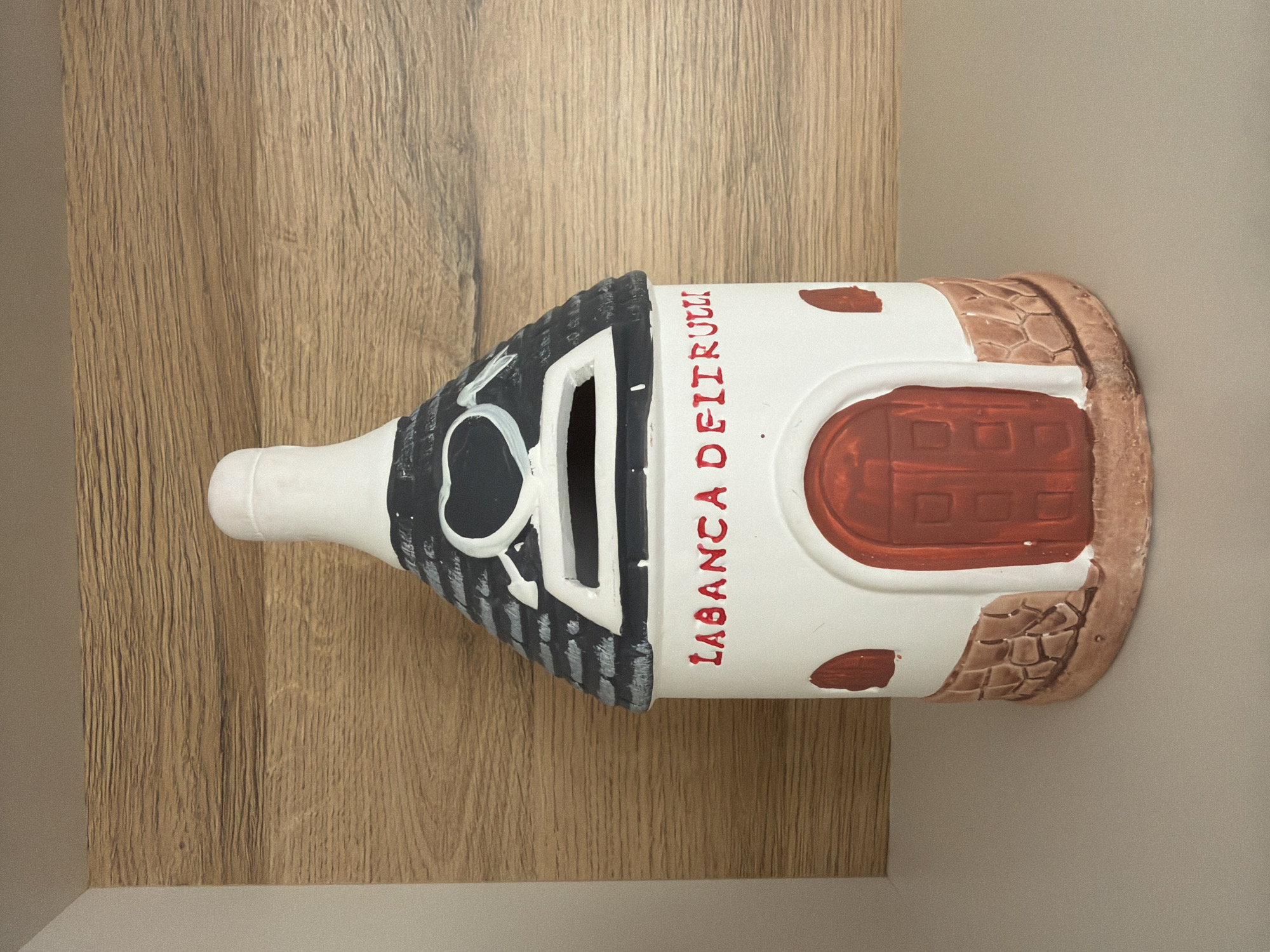 This is a medium shot, eye-level photograph of a white ceramic piggy bank shaped like a house with a dark gray, shingled roof. The roof is adorned with a white heart with an arrow through it and a white opening for inserting coins. The house portion of the piggy bank has red lettering that reads "LABANCA DEI FIURULLI". There is a red arched doorway with a darker red door featuring square indentations. To the left and right of the door are small, arched red windows. The base of the piggy bank is decorated with faux brown stones. The piggy bank sits on a light gray surface, and behind it is a wooden paneled wall.
This object does not have a specific location in the city of Pazardzhik, Bulgaria. It is a decorative item.