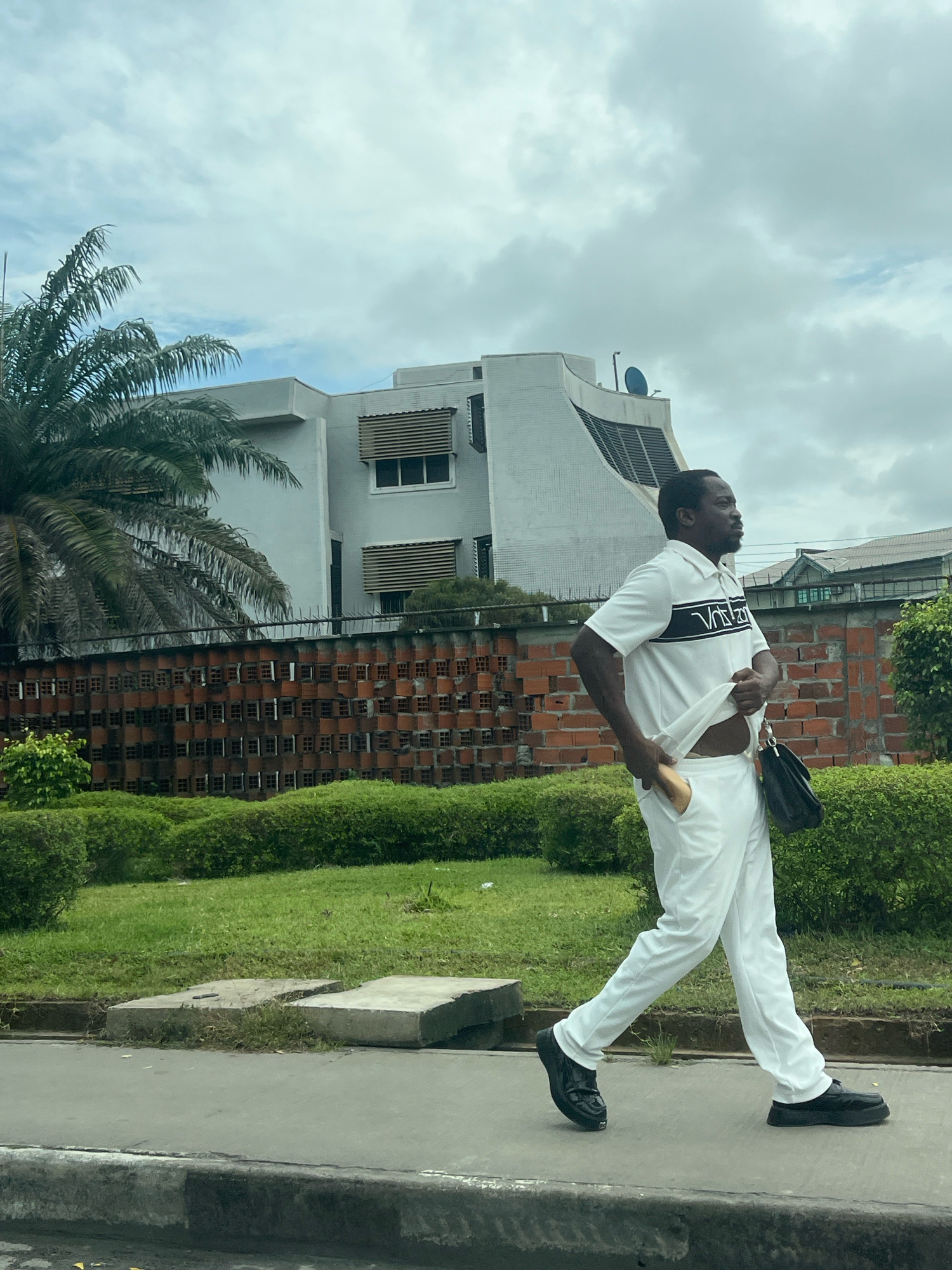 A man is walking on a sidewalk in Lekki Phase I, Nigeria. He is wearing a white polo shirt with black stripes and a white logo on the chest, and white pants. He is holding his shirt up with his right hand, revealing his midriff. He has a black bag slung over his left shoulder. He is wearing black loafers. Behind him is a brick wall with a patterned design, a well-maintained green hedge, and a modern white building with a curved facade. A palm tree is visible to the left of the building. The sky is cloudy. The sidewalk is made of concrete, and there is a strip of grass and concrete slabs beside it.