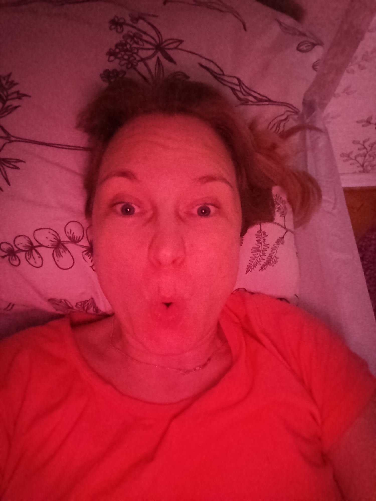 A woman is lying on a bed with patterned bedding. She is wearing a red shirt and has blonde hair. Her face is close to the camera and she has her mouth open in a surprised or playful expression. The lighting in the room is red or pink-toned. The exact location of the photo is unclear.