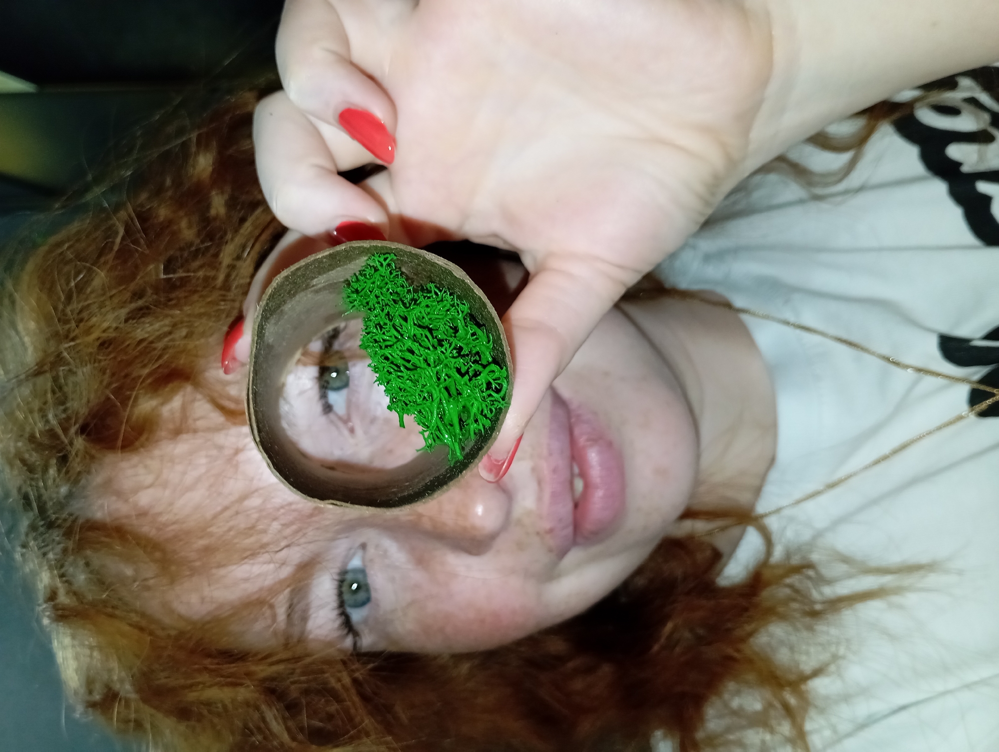 An angled, close-up portrait shows a person with red or auburn hair and a freckled face, looking towards the viewer. Their head is tilted, presenting their face sideways. One hand, adorned with bright red nail polish, holds an empty cardboard tube directly in front of their face. Inside the tube, a vibrant cluster of bright green, textured artificial moss or similar material is visible. Through the tube's opening, one of the person's green-blue eyes is clearly seen, making direct eye contact, while the other eye and pink lips are partially visible below the tube. A thin golden chain necklace hangs on their chest, which is covered by a white garment featuring abstract black patterns. The background on the left is dark and indistinct, contrasting with the lighter, patterned fabric on the right, suggesting a casual indoor setting. The image portrays a creative or playful pose, using the tube and moss as a photographic prop. There is no visible text.