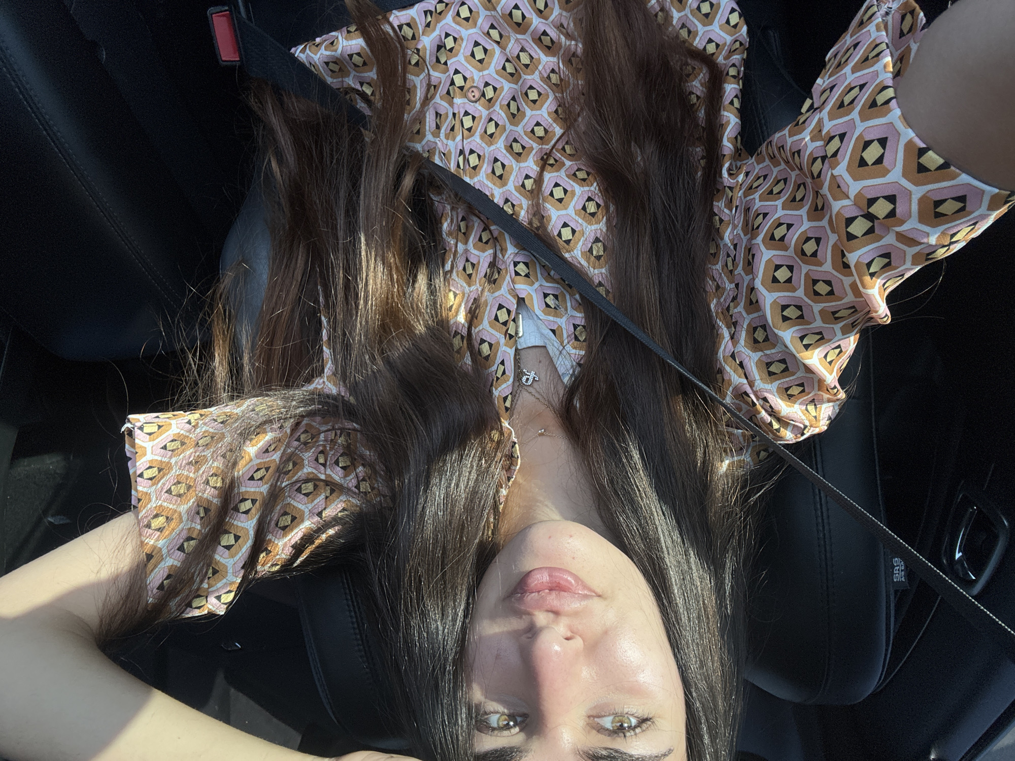 A young woman with long, dark hair and light-colored eyes is seated in the driver's seat of a car. She is wearing a collared, short-sleeved shirt with a geometric pattern in shades of pink, orange, yellow, and black. A silver necklace with a small pendant is visible around her neck. The car's interior is dark, with black leather seats and a seatbelt diagonally across her chest. Sunlight illuminates her face and hair, casting shadows. The scene appears to be in Deerfield Beach, Florida, United States of America.