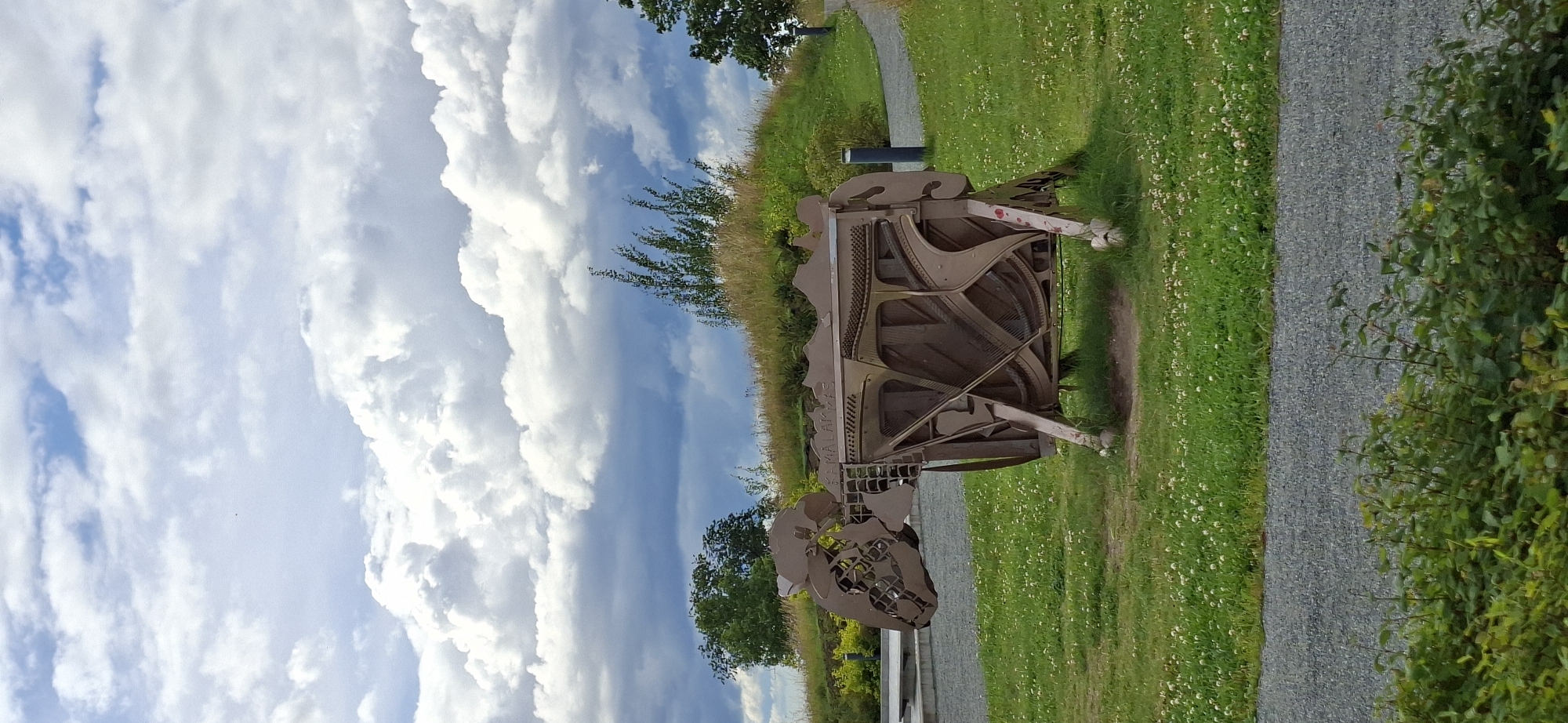 An outdoor sculpture of a sheep made from scrap metal, possibly old piano parts, stands on a grassy hill under a cloudy sky. The sculpture is positioned in the center of the frame, facing left, with its head lowered. The background features a green hill with trees and a paved path winding through it. The foreground is a grassy area with scattered white flowers, bordered by a gravel path and dense green foliage at the bottom of the image. The sculpture is located in Rakvere, Estonia, specifically at the Rakvere Castle grounds, where it is part of the art installations. The sculpture appears to be a static exhibit, with no visible people or animals interacting with it. The environment suggests a public park or historical site.