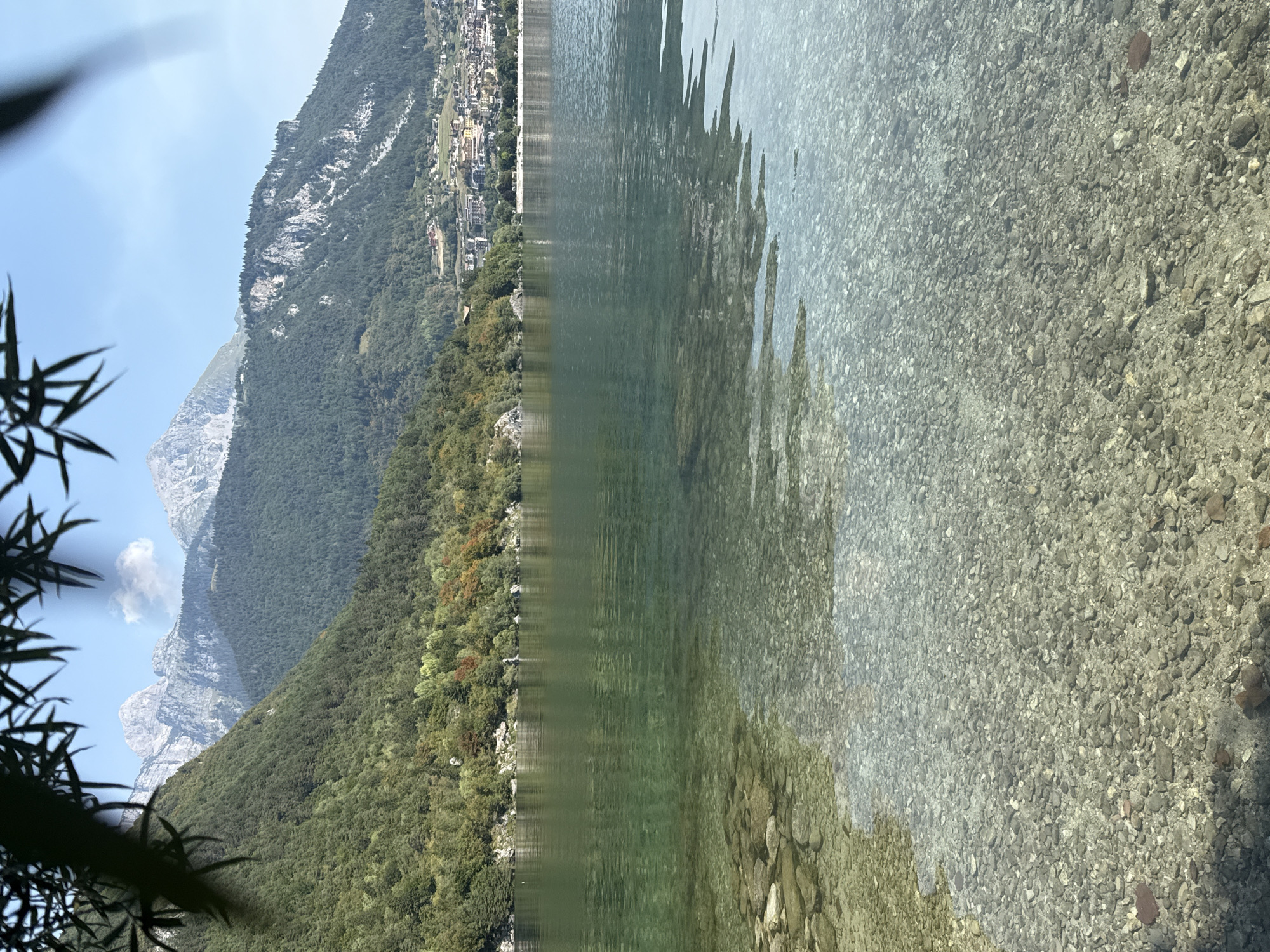 The image shows a clear, shallow lake with a rocky bottom, surrounded by forested mountains and a distant town. The water is a translucent turquoise color, with ripples on the surface. The lakebed is covered in smooth, multi-colored pebbles of various sizes. In the background, a densely wooded mountain slopes down towards the lake. Beyond the mountain, a town with buildings of various colors is visible. The sky is a clear blue with a few scattered white clouds. The image appears to be taken on a sunny day. The foreground is dominated by the lake's clear water and rocky bottom, while the background features a dramatic mountain landscape and a small town. The overall impression is one of natural beauty and tranquility.