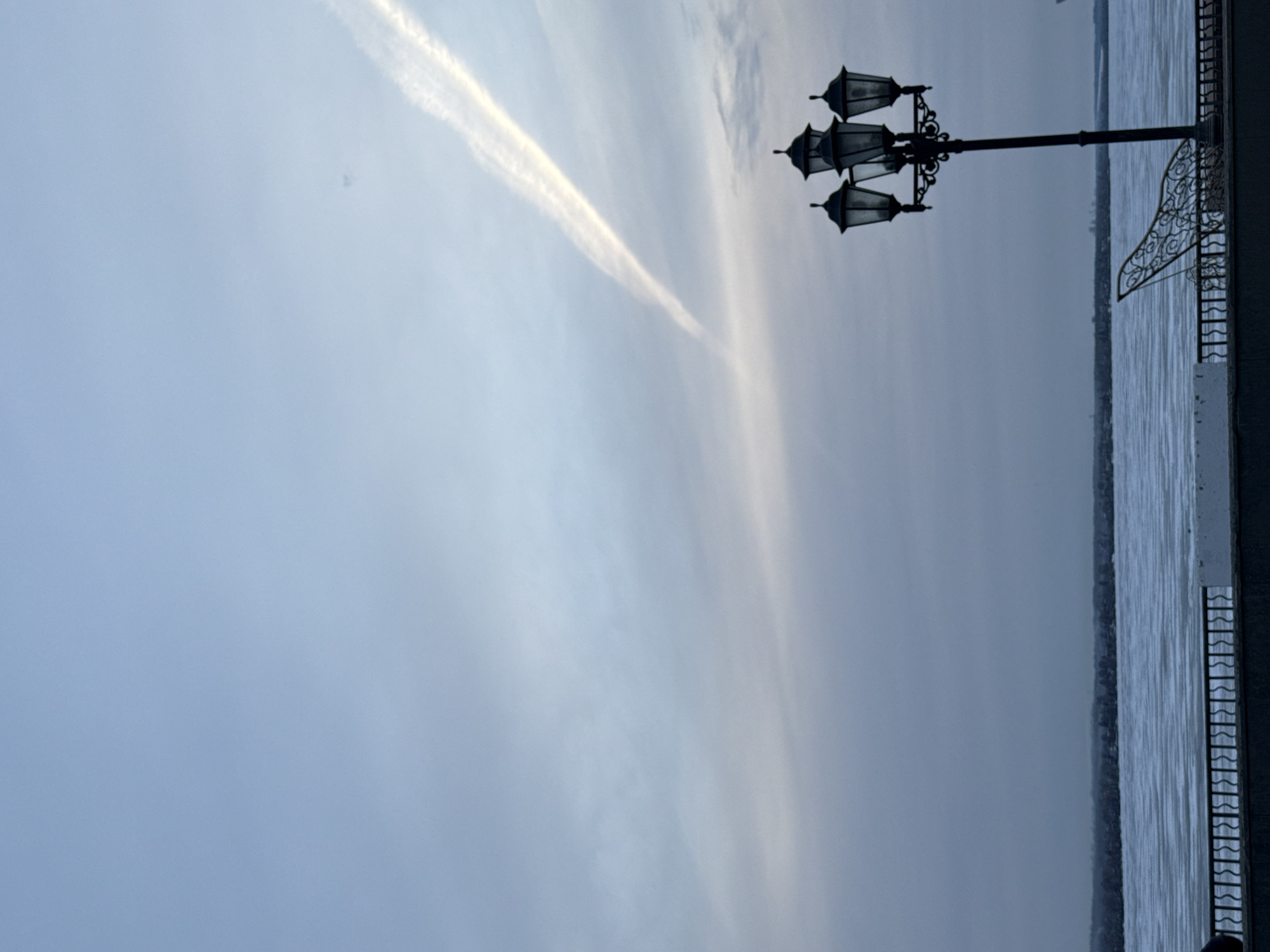 The image shows a street scene in Votkinsk, Russia. The sky is overcast with a prominent contrail streaking across the upper portion of the frame. A decorative lamp post with five globes stands on the right side of the image, near what appears to be a waterfront area. A metal railing runs along the edge of the waterfront. The water is visible, suggesting a river or lake. The overall tone is muted due to the overcast conditions.