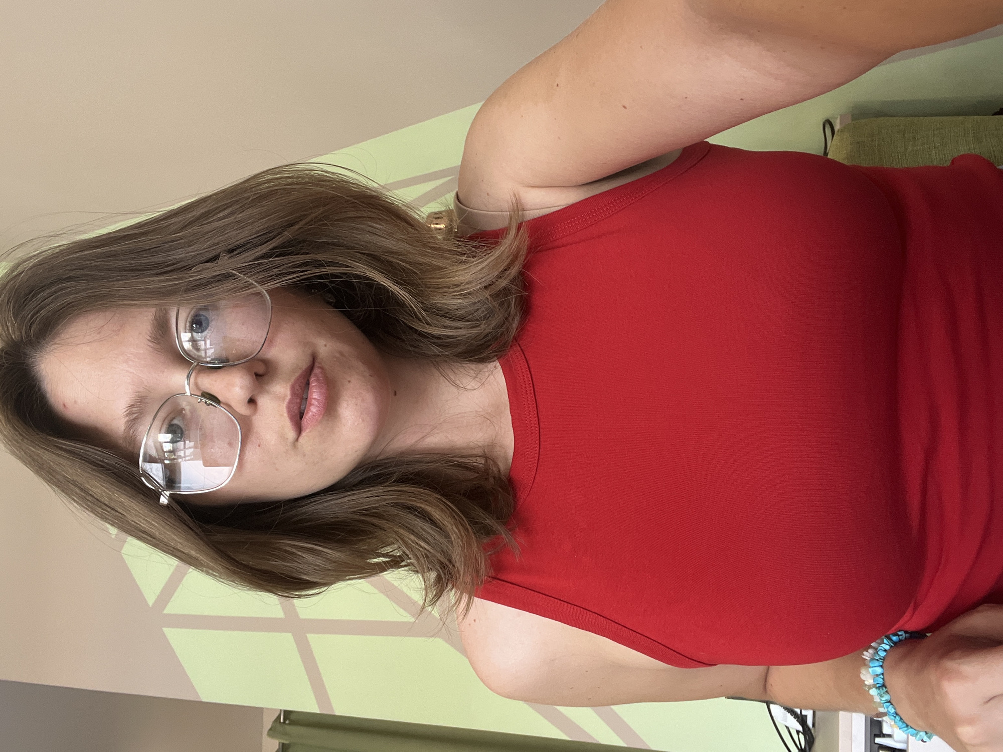 A woman with light brown hair and silver-rimmed glasses is wearing a red tank top. She has a blue beaded bracelet on her wrist. The background features a light green and beige wall with geometric shapes. The location is not provided in the image.