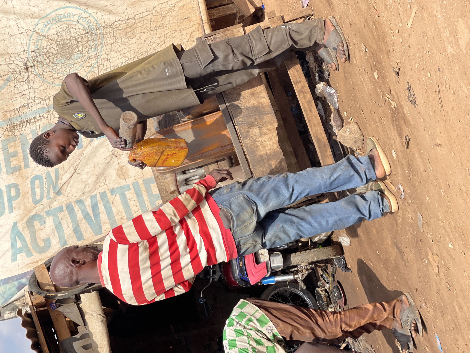 Two men stand outside near an old, weathered sign in a dusty urban area. One man wears a red and white striped shirt and blue jeans. He stands near a parked motorcycle. The second man, in a green uniform, pours liquid from one container into another. The location is likely Abuja, Nigeria.