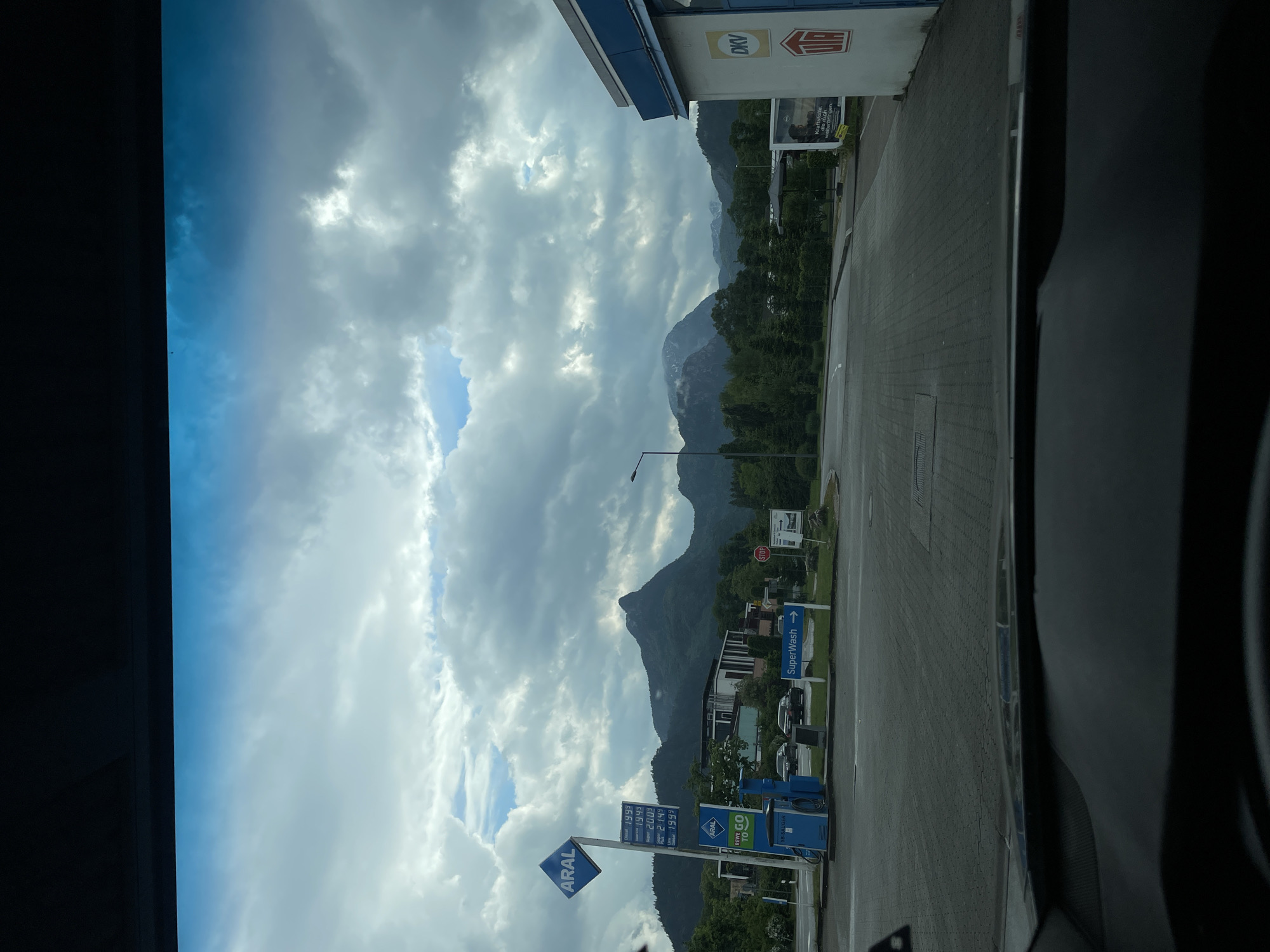 A sideways view from inside a vehicle, possibly a car, shows a roadside scene in Unknown, Unknown. The immediate foreground is dominated by the vehicle's interior, with a dark dashboard visible. Outside, the scene is split between a petrol station and a scenic mountainous landscape.
On the right, a petrol station is clearly recognizable with signage featuring logos for "ARAL" and "DKV." There are also signs for a "SuperWash." Price boards are visible, showing prices per liter for various fuels. The ground is paved, appearing to be a parking or refueling area.
In the background, mountains rise against a sky filled with clouds. The landscape is green, indicating trees and vegetation. The sky is varied, showing patches of blue sky mixed with thick, grey clouds. The light appears somewhat diffused, suggesting an overcast day. There are no people visible in the shot. The view is partially obstructed by the roadside infrastructure.
