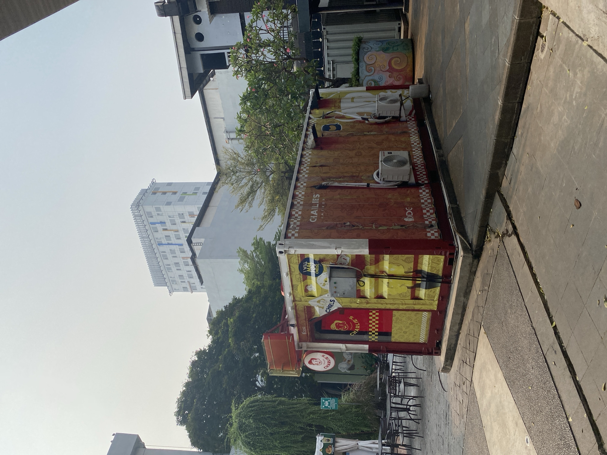 In North Jakarta, Indonesia, a red and yellow shipping container repurposed as a food stall, "CHARLIE'S," is situated on a paved outdoor area. The stall is decorated with cartoon graphics and the "CHARLIE'S" logo. Two outdoor air conditioning units are mounted on its exterior. In front of the stall, tables and chairs are set up on a patterned tiled surface. Behind the stall, a large, leafy tree and a tall building with a white facade and colorful windows dominate the background. The sky is overcast with a hazy, light grey hue.