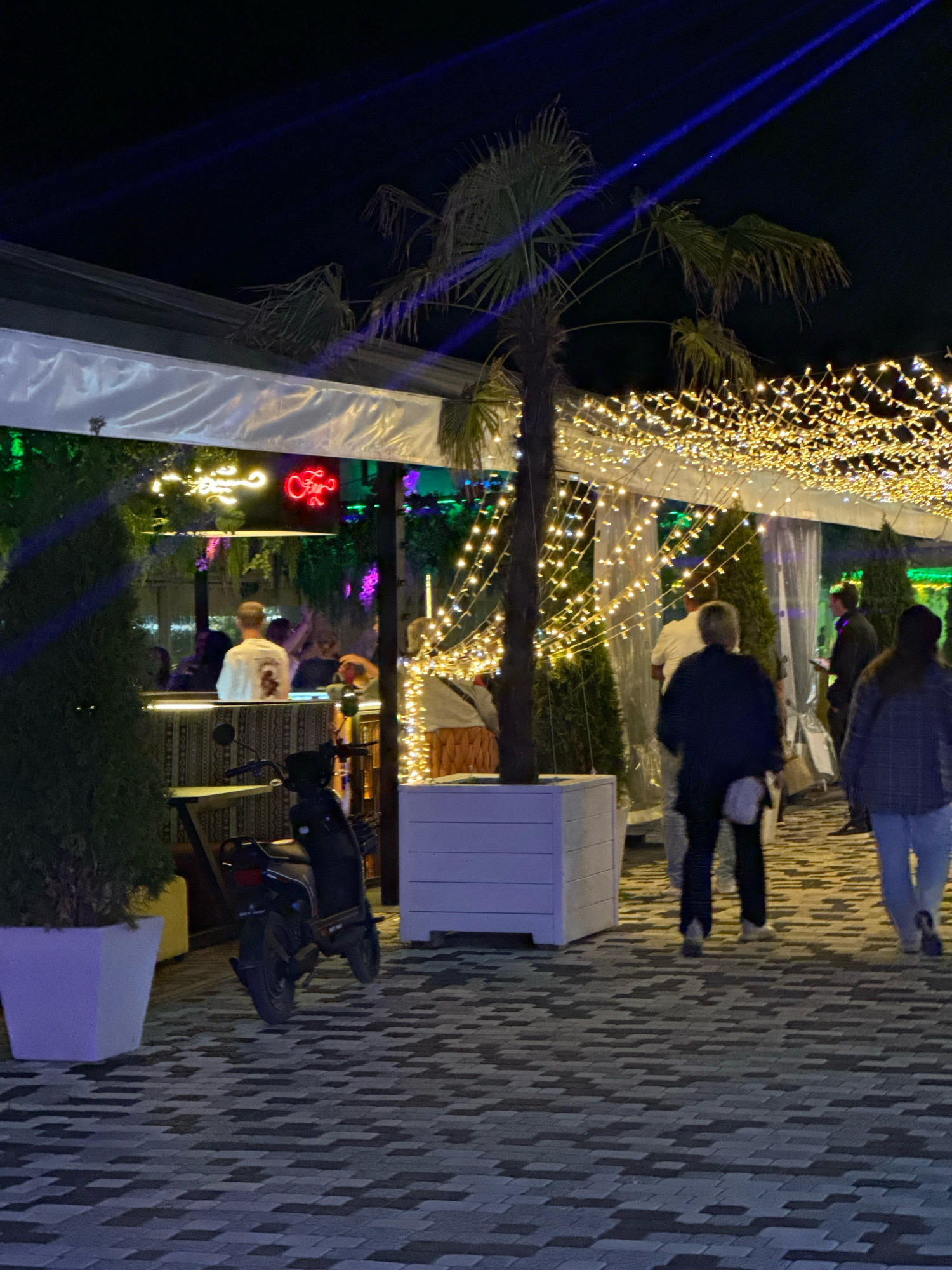 The image depicts an outdoor seating area at night, possibly a cafe or restaurant, with blue light beams projected from above onto the dark sky. String lights in warm yellow tones are draped across the ceiling. In the foreground, a paved walkway is visible, with two cypress trees in white planters on the left and a white planter box on the right. A dark scooter is parked to the left of the walkway. Several people are walking and standing in the background, some seated at tables under a covered area illuminated by neon signs. One person, facing away from the camera, is wearing a white shirt with a graphic design. The overall atmosphere suggests a social gathering or dining experience in a nighttime urban or resort setting.