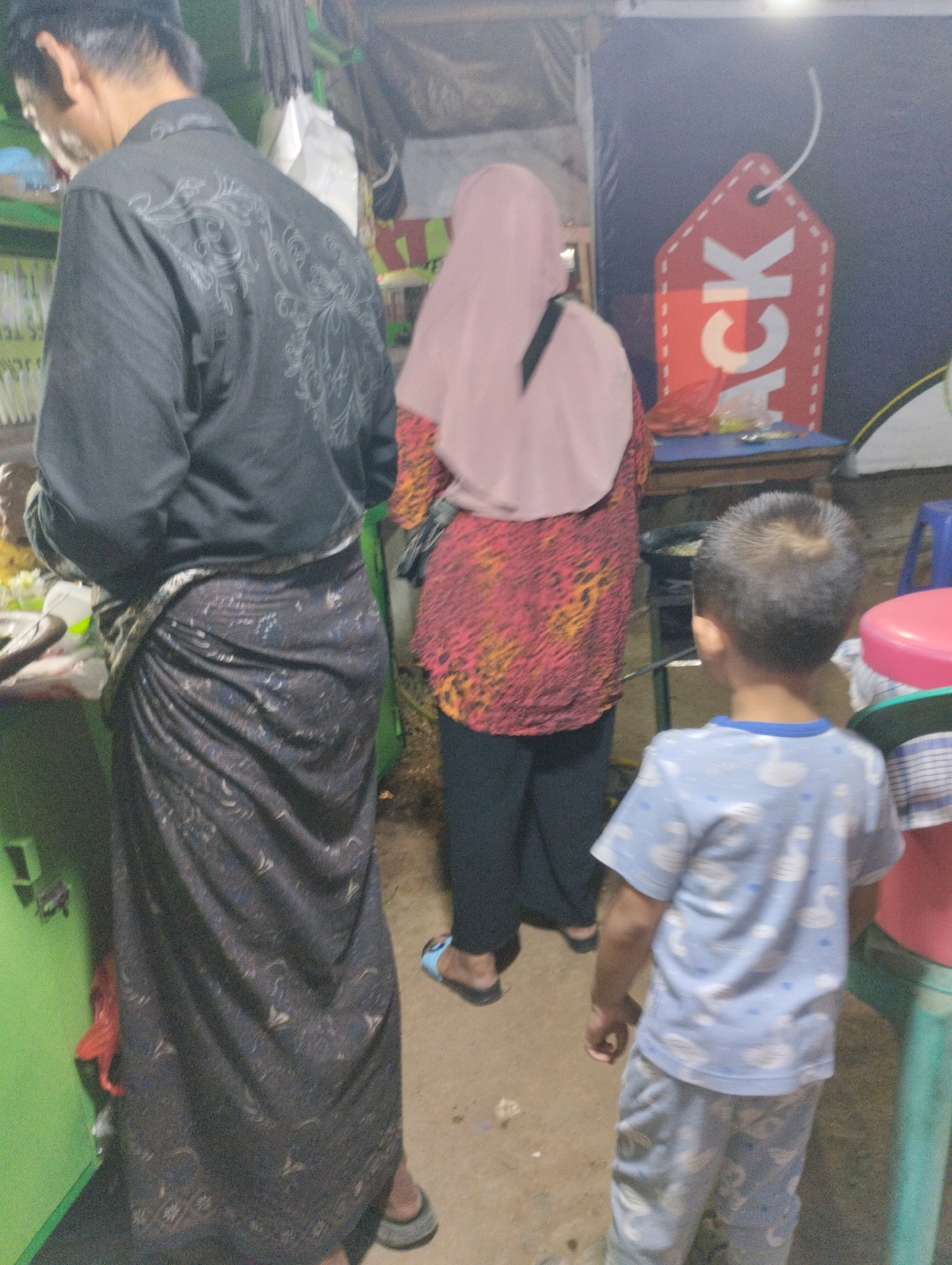 The image depicts a street scene in Bekasi, Indonesia, likely an outdoor market or food stall area. In the foreground, a man wearing a black patterned shirt and a dark sarong stands to the left. To his right, a woman in a pink hijab and a red and yellow patterned top is partially visible. A young boy in a light blue patterned shirt and pants stands facing away from the camera, looking towards a table with food items. Behind the people, a red tag with the word "ACK" is displayed prominently against a dark background. The environment appears to be cluttered with various items, including plastic bags, containers, and stools. The lighting suggests it is either dusk or nighttime, with artificial light sources illuminating the scene.