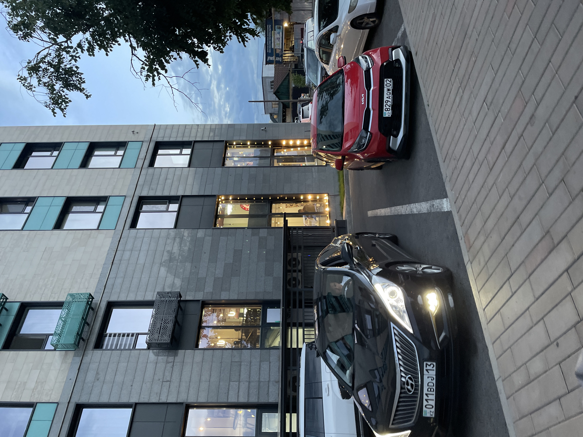 The image shows a residential building in Almaty, Kazakhstan, with a modern facade featuring a mix of grey stone and turquoise accents. Several cars are parked in front of the building on a paved area. A black Hyundai sedan is in the foreground, with its headlights on, and a person is visible in the driver's seat. To the right, a red Kia hatchback is parked, also with its license plate visible. Other vehicles, including a white SUV, are also present. The sky is overcast, suggesting it might be late afternoon or early evening. The building's windows are illuminated, some with decorative string lights, indicating activity within. The scene appears to be a typical urban setting in Almaty.