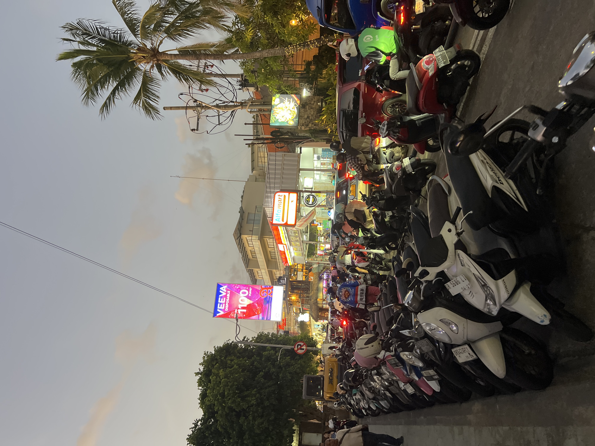 The image depicts a street scene in Kuta, Indonesia, at dusk. A long row of parked scooters and motorcycles lines the left side of the street. The scooters are predominantly light-colored, with a few darker models interspersed. Several people are visible walking or on motorcycles. On the right side of the street, traffic is congested, with cars and more scooters. Buildings with illuminated signs are visible in the background, including an "Alfamart" store and a large digital billboard advertising "VEEVA DISPOSABLE 1100 PUFFS". A palm tree and electrical wires are also prominent features of the urban landscape. The lighting suggests that it is either early evening or late afternoon.