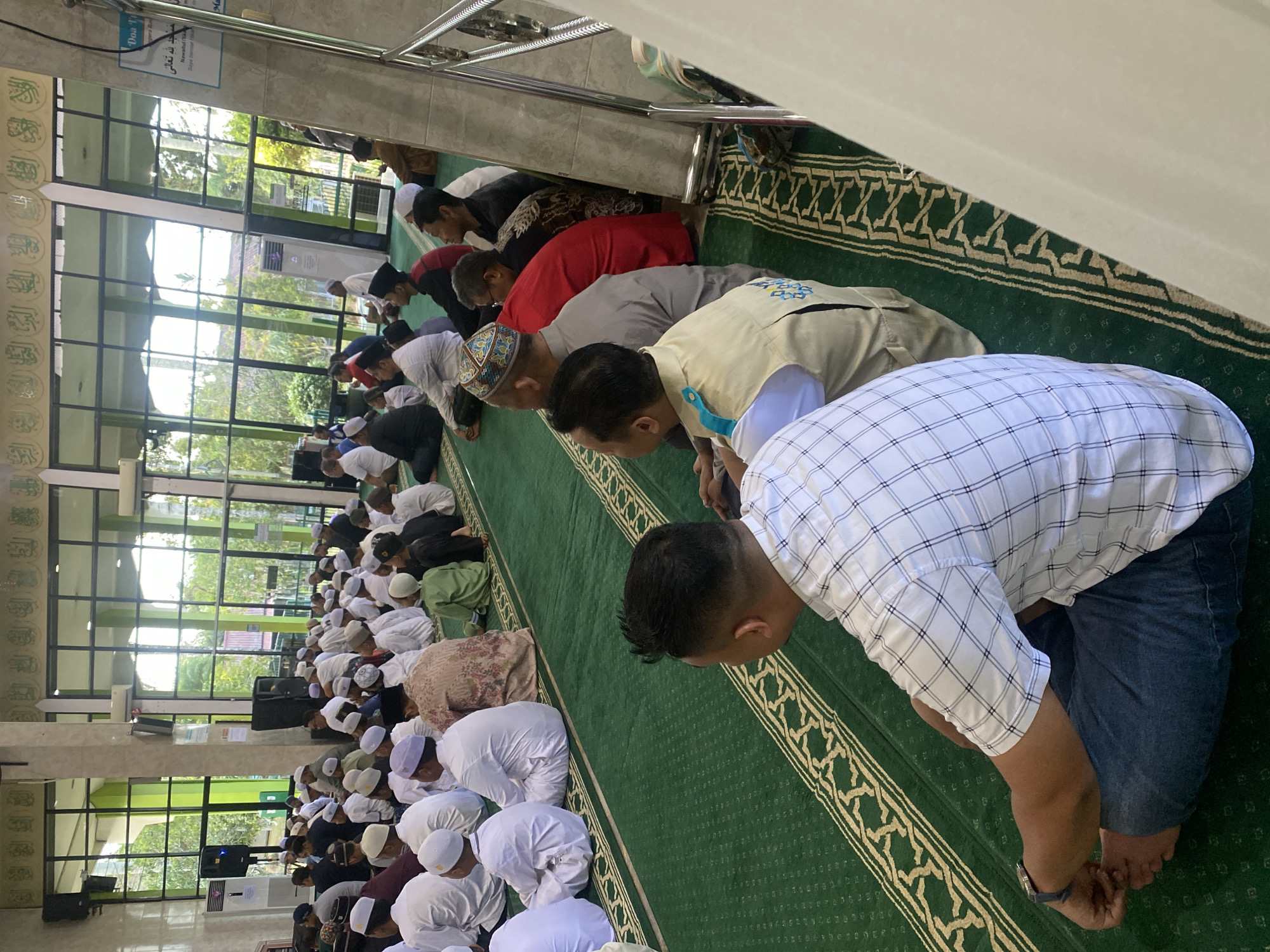 A group of men, likely performing congregational prayer, are seated on a green carpeted floor inside a mosque. The men are dressed in various attire, including white robes and skullcaps, some in casual clothing. The mosque features large windows with a view of trees and an overcast sky. The interior includes air conditioning units, speakers, and decorative calligraphy on the upper walls. A sign with Arabic script is visible on a column. In the foreground, a man in a checkered shirt and jeans is seated, facing away from the camera. The scene is set in the city of Banjarbaru, Indonesia.