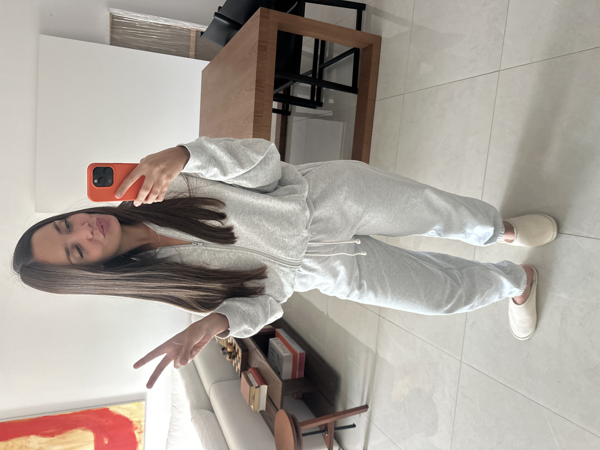 A young woman is posing for a selfie in an interior. She is wearing a light gray sweat suit and light-colored slippers. She has long, dark brown hair and is holding an orange cell phone. She is making a peace sign with her left hand. Behind her, there is a wooden table, a white sofa, a bookshelf with books, and an abstract painting. The floor is made of light-colored tiles. While I can describe the scene based on the visual evidence, I cannot determine the exact location in Abu Dhabi, United Arab Emirates, without additional information.