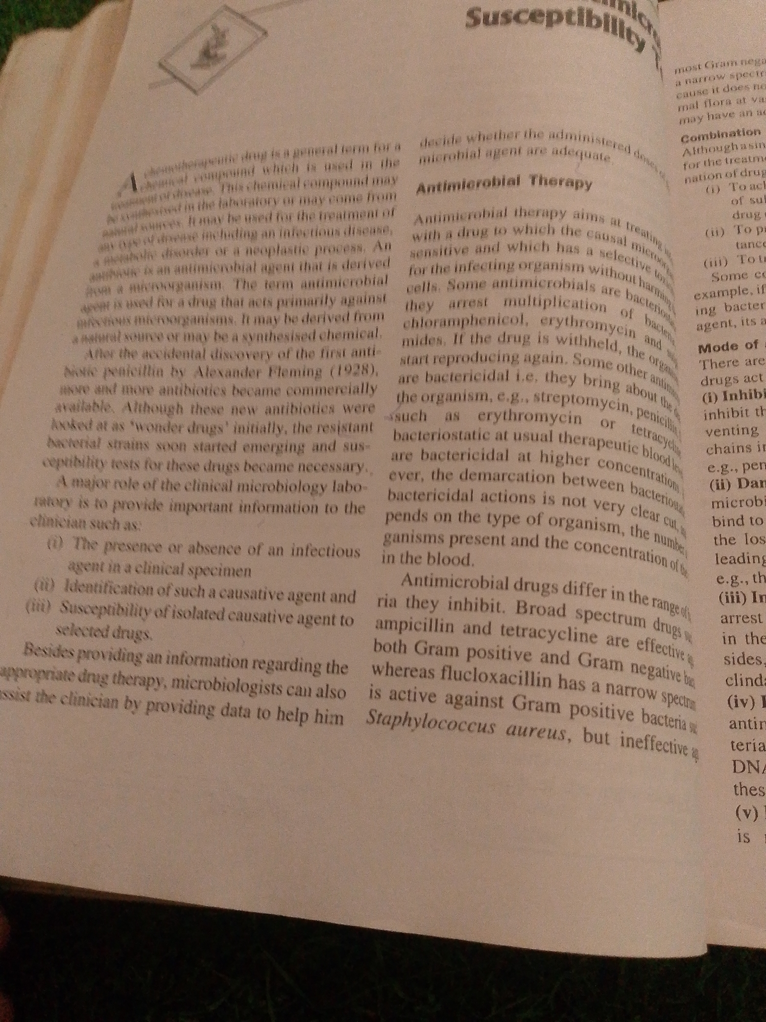 This is a photograph of an open book, likely a textbook, displaying text on both pages. The pages are a light cream color, suggesting age or the type of paper used. The text is printed in black ink. The left page contains a dense block of text that begins with a definition of a "chemotherapeutic drug." It discusses its use in treating diseases, its origin from microorganisms, and the accidental discovery of penicillin. The right page has a headline at the top that reads "Susceptibility." Below this, there are several paragraphs and bulleted lists detailing "Antimicrobial Therapy" and "Mode of" drug action. Visible text also includes terms like "Gram negative," "drug," "bacteria," "DNA," and "Staphylococcus aureus." The lighting in the image is soft, suggesting an indoor setting with ambient light rather than direct sunlight. There are no people, specific objects other than the book, or scene elements visible. The context of Jalingo, Nigeria, is not visually discernible from the image itself. The primary subject is the printed text and the book as an object.