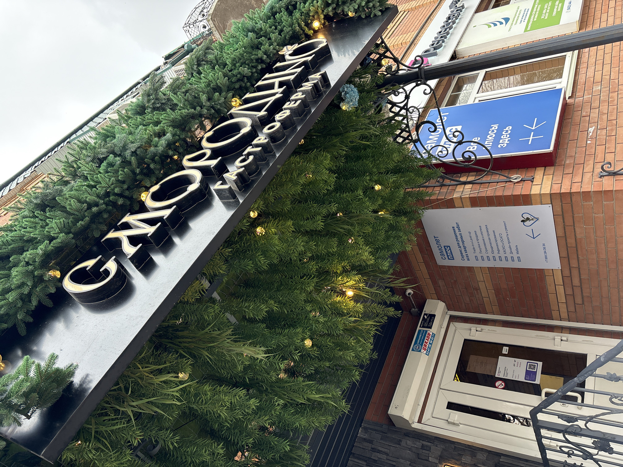 The image shows the exterior of a building in Ulyanovsk, Russia. A black sign with the gold-colored Cyrillic words "СМОРОДИНА ГАСТРОФЕРИА" is prominently displayed, surrounded by green foliage and small lights. To the right of the sign, a blue sign with the Cyrillic word "ПЛЮСЫ ЗДЕСЬ" is visible above a smaller white sign with information in Cyrillic. Below the white sign, a white door with glass panels and a QR code sticker is visible. A security camera is mounted above the door.