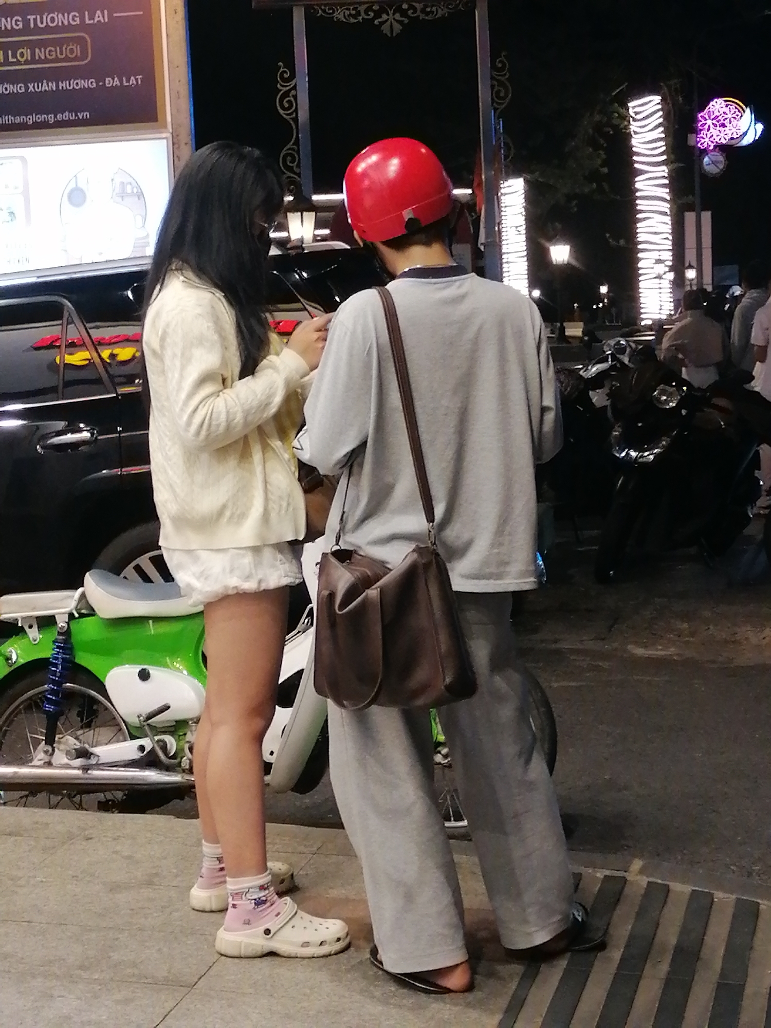 Two individuals are standing next to a motorcycle and a black car at night. The person on the left appears to be a woman with long black hair, wearing a cream-colored cardigan over a white dress and pink socks with white clogs. The person on the right, facing away from the camera, is wearing a red helmet, a grey t-shirt, and grey pants. A brown messenger bag is slung across their body. The setting is outdoors, likely a street or sidewalk, with streetlights and illuminated signs visible in the background. Motorcycles are parked nearby. The lighting suggests it is nighttime. There is visible text on a sign in the upper left corner of the image, which appears to be in Vietnamese. The overall mood seems casual and everyday.