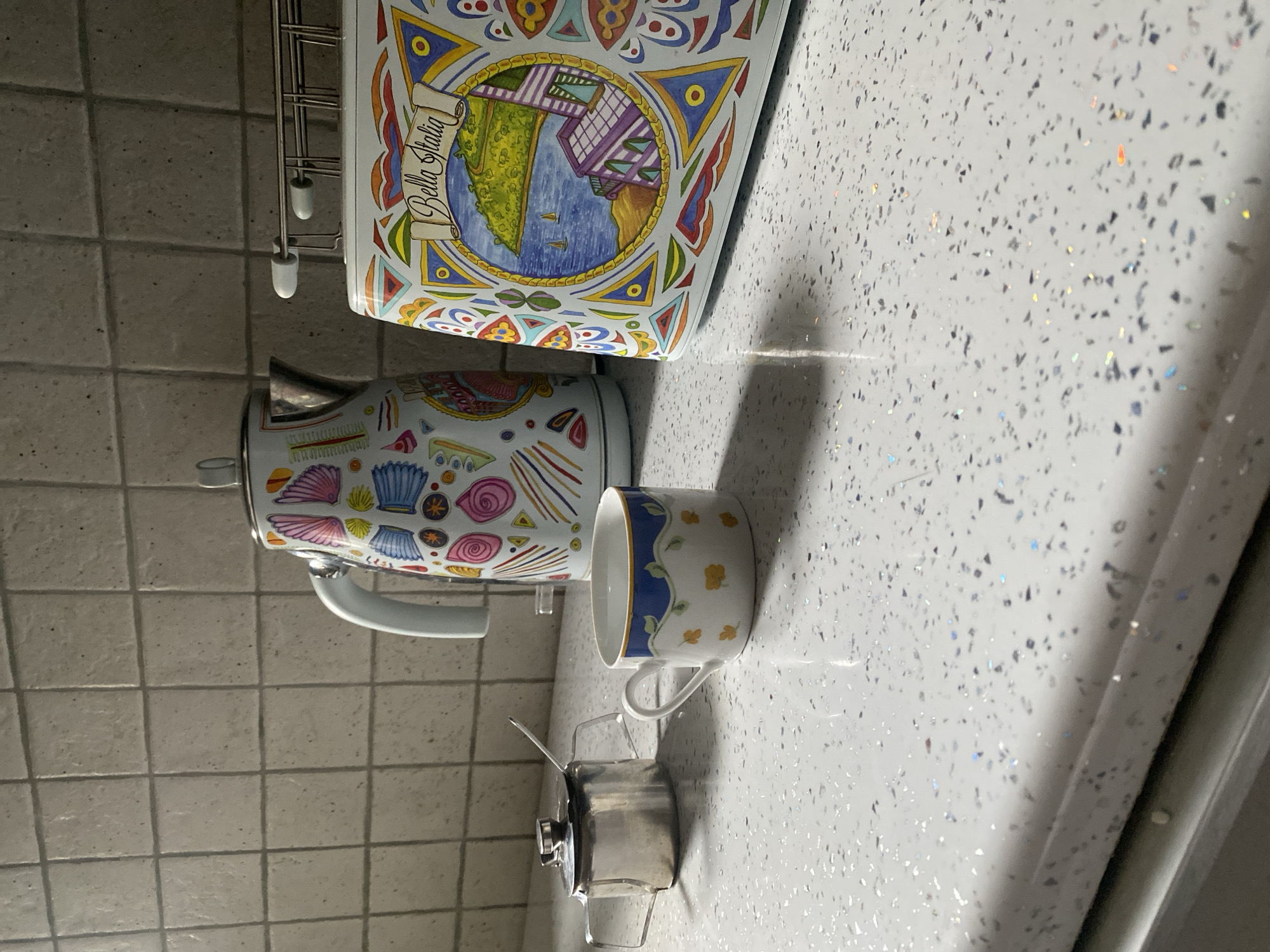 The image depicts a kitchen counter with several decorative items. In the foreground, a white ceramic cup with blue and yellow floral designs sits next to a small, silver metal container with a lid and a spoon. Behind these, a white electric kettle is visible, adorned with colorful, whimsical patterns, including stylized shells and geometric shapes. To the right of the kettle, a toaster with a similar vibrant, artistic design, featuring a scenic Italian coastal village depicted on its front, is present. The countertop itself is white with a speckled pattern, reflecting light. The background consists of light-colored square tiles on the wall. The scene is lit by a light source from the left, casting shadows on the counter. There are no people present in the image. The specific location in the city of Wrocław, Poland, cannot be determined from the visual information provided in the image.