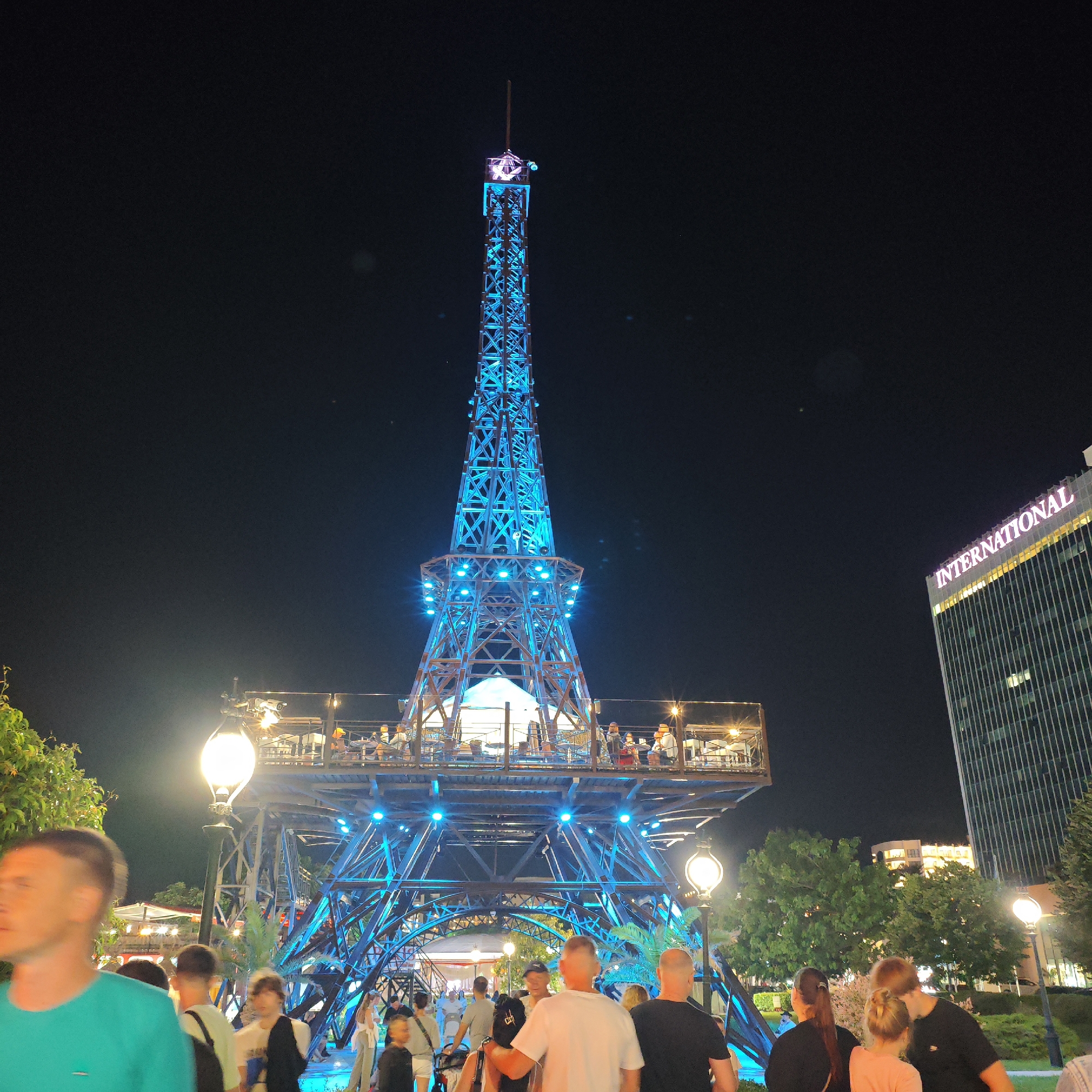A brightly lit replica of the Eiffel Tower, illuminated with blue lights, stands as the central feature of a public area at night. The tower has multiple levels with people dining on the upper platforms. In the foreground, several individuals are walking, with some facing the tower and others walking away. To the right of the tower, a modern, tall building with the word "INTERNATIONAL" illuminated in pink on its facade is visible. The surrounding area includes trees and streetlights, suggesting an urban entertainment district. The scene appears to be in the city of Varna, Bulgaria, likely at the resort complex.