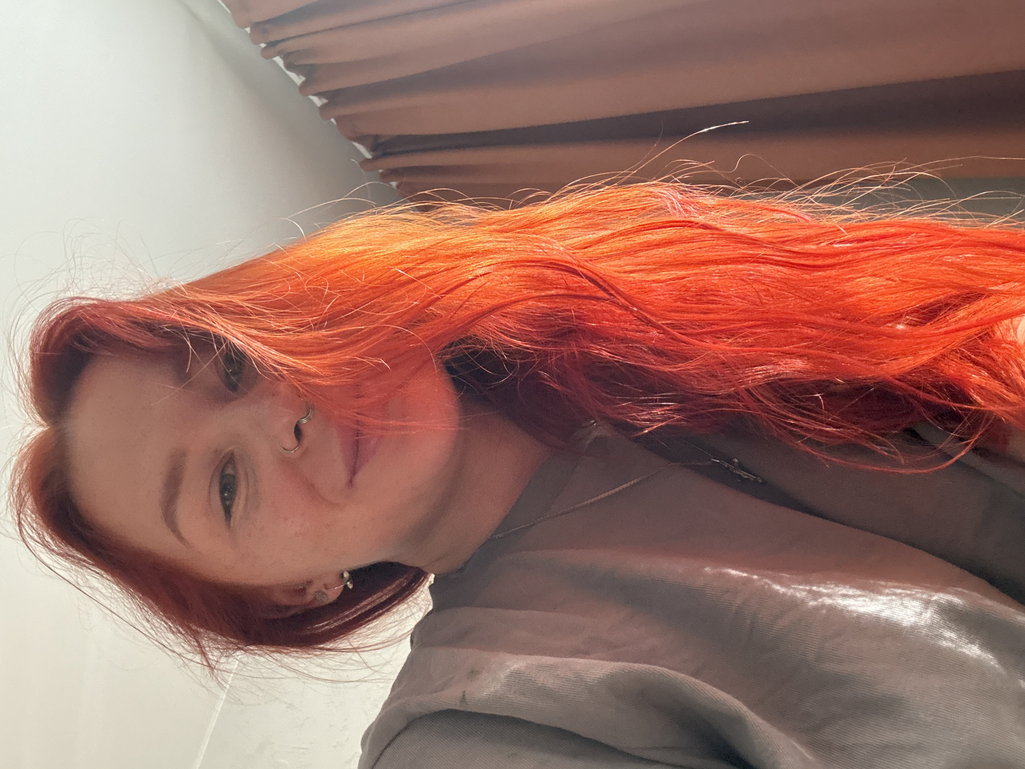 The image depicts a person with bright red, long hair, a nose ring, and freckles. They are wearing a grey shirt and a delicate necklace. The background shows part of a wooden structure, potentially blinds or a headboard. The location cannot be determined from the image, so it cannot be stated if the image was taken in the city of Rostov-on-Don, Russia.