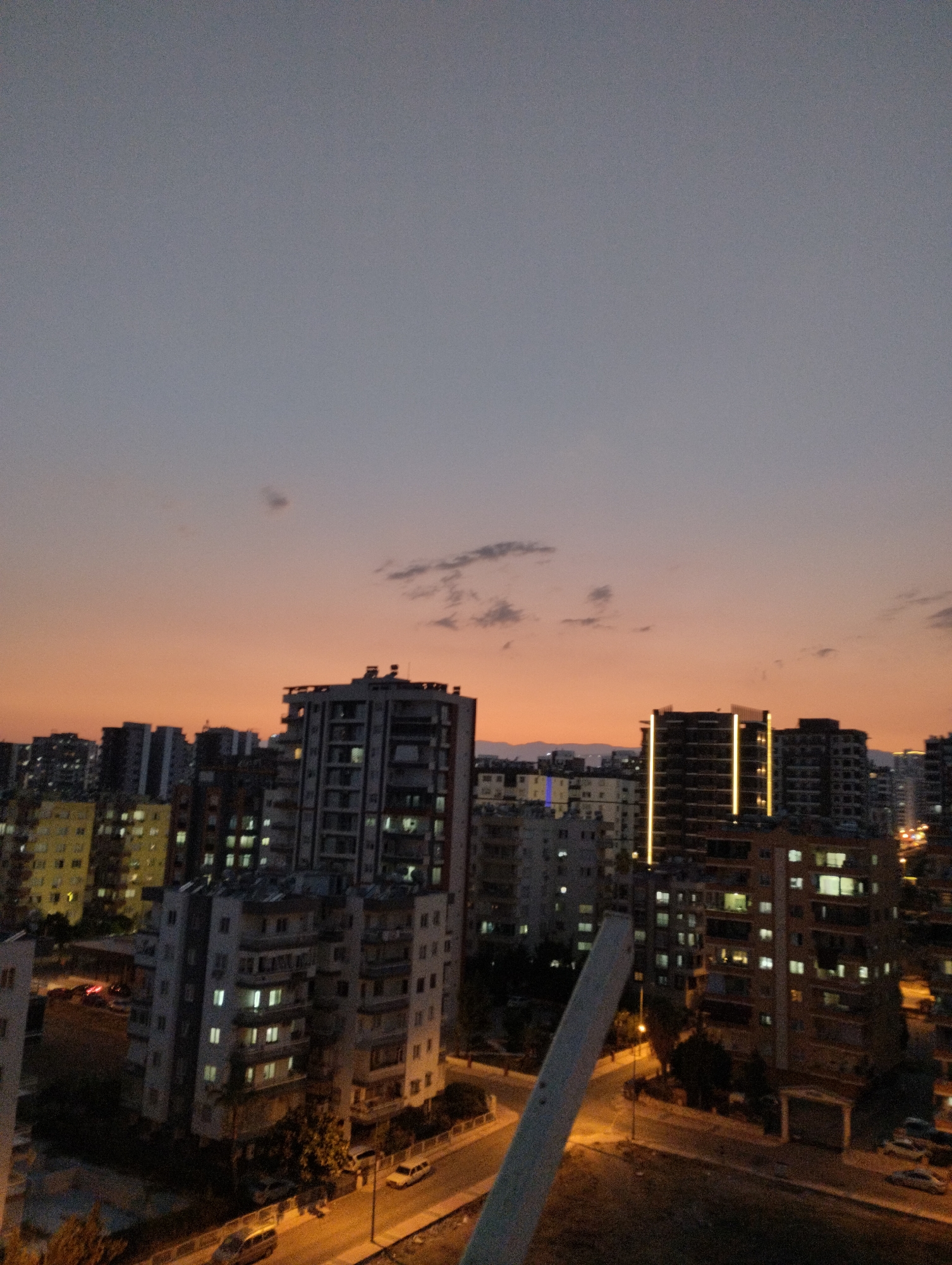 The image depicts a cityscape at dusk, likely in Mezitli, Turkey, given the architectural style of the buildings. The sky displays a gradient of colors, transitioning from a pale gray at the top to a warm orange hue near the horizon, indicating the setting sun. Scattered clouds are visible in the sky.
The foreground is dominated by a series of multi-story apartment buildings. Many of these buildings have illuminated windows, suggesting that it is evening and residents are home. Some buildings are taller and more modern, featuring prominent vertical lighting elements. Others appear older and less illuminated.
Below the buildings, a street is visible, illuminated by streetlights. Several vehicles, including cars and possibly a van, are parked along the street or driving on it. The presence of cars and apartment blocks points to a residential and urban environment. Trees and landscaping are sparsely distributed between buildings and along the street. The overall impression is of a typical urban scene as day turns into night.