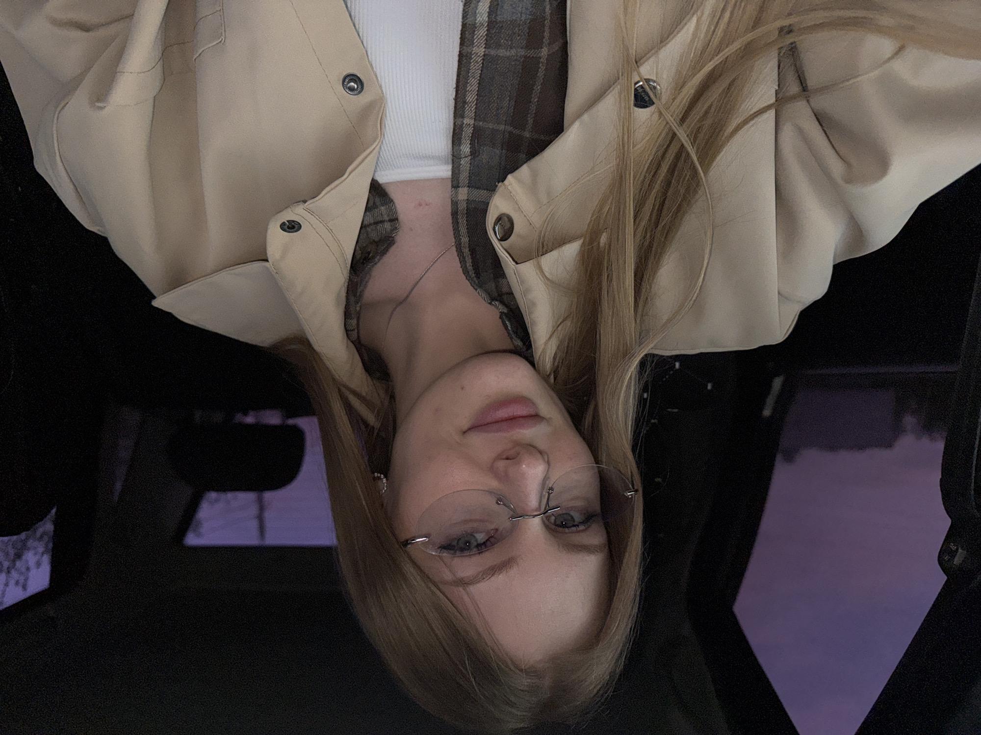 A blonde, fair-skinned woman wearing rimless glasses and layered clothing (a beige jacket, a plaid shirt, and a white undershirt) is pictured in an upside-down photograph. The woman appears to be in a car, based on the interior car parts visible in the dark background, including a rearview mirror and a window frame reflecting a purplish-blue twilight sky. There is no indication of a location in the city of Perm, Russia.