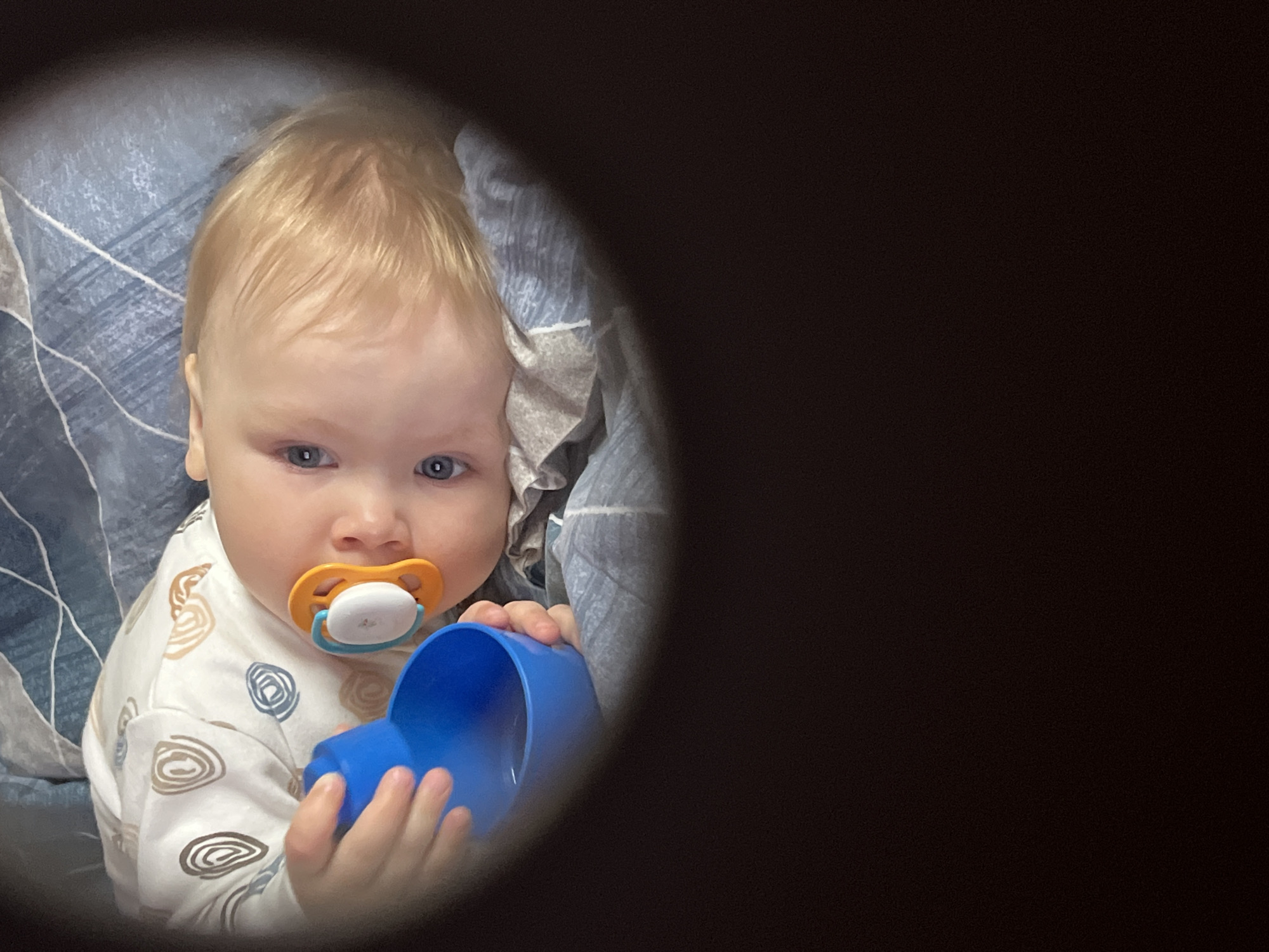 A baby with blonde hair and blue eyes is holding a blue plastic cup and has a pacifier in its mouth. The baby is wearing a white onesie with brown swirl patterns. The background includes a blue and gray patterned blanket. The photograph appears to be taken through a dark hole, obscuring a large portion of the right side of the frame. It's not possible to confirm the location as Chaikovsky, Russia, without further information.