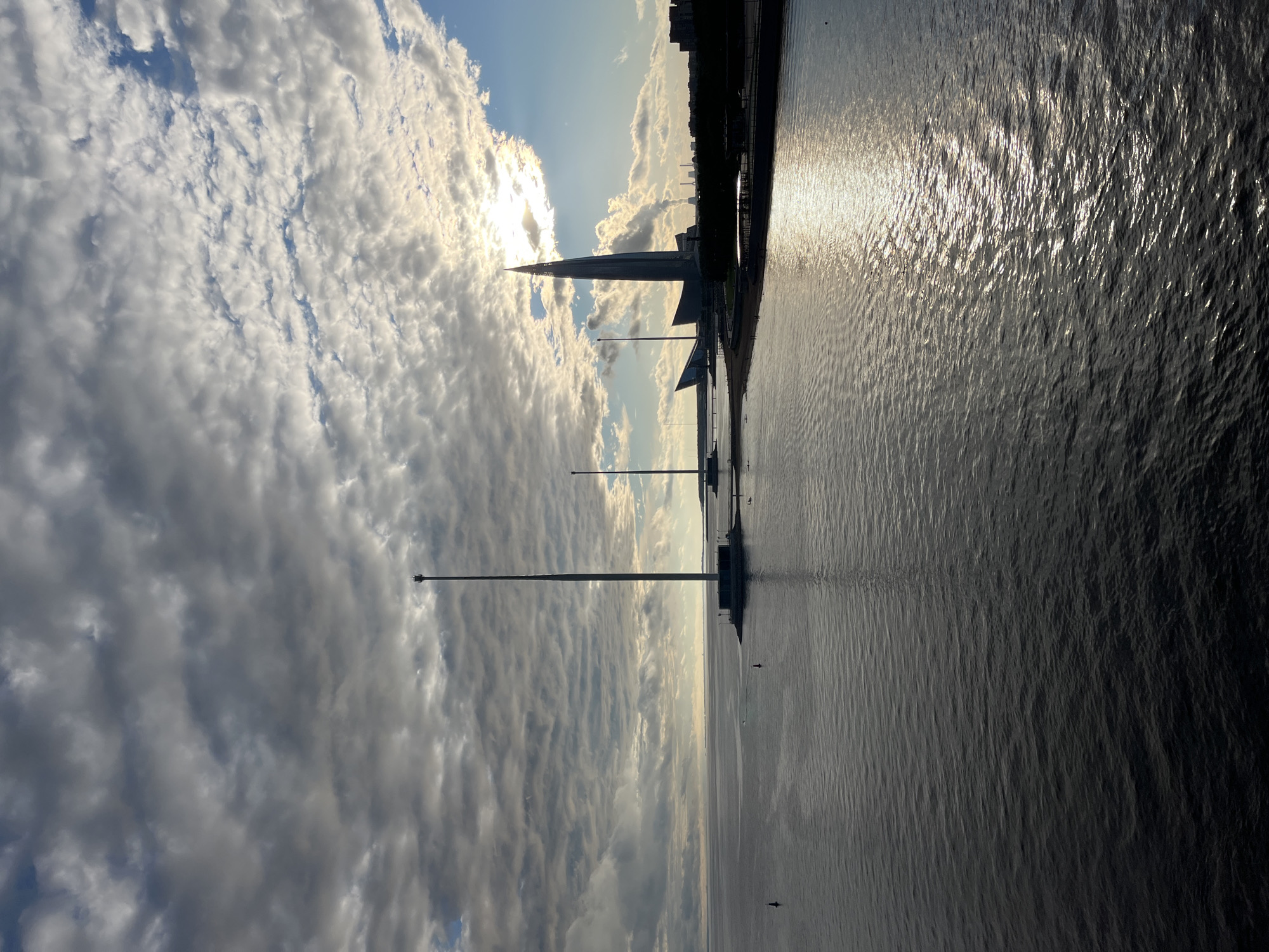 This is a landscape photograph of a waterfront scene in Saint Petersburg, Russia, taken during what appears to be late afternoon or early evening. The image is oriented vertically.
The foreground is dominated by the dark, rippling surface of a body of water, likely a gulf or bay, reflecting the sunlight which creates a bright, shimmering path across the water. The water is mostly calm, with small waves creating texture.
In the mid-ground, there is a distinct, futuristic-looking skyscraper with a pointed top, silhouetted against the bright sky. This is the Lakhta Center. To the right of the skyscraper, a cityscape is visible along the horizon, appearing as dark shapes against the sky. Several tall, slender poles, likely for lighting or flags, are spaced along the waterfront. A few small boats or vessels are visible on the water.
The sky is filled with dramatic, textured clouds. Some are bright white and illuminated by the sun, while others are darker and more shadowed, creating a dynamic contrast. The sun itself is partially obscured by clouds, casting a bright glow.
There is no visible text in the image. The overall mood is serene and somewhat dramatic, due to the contrast between the light and dark elements, the striking architecture, and the expansive sky and water. The scene captures a sense of urban grandeur meeting natural elements.