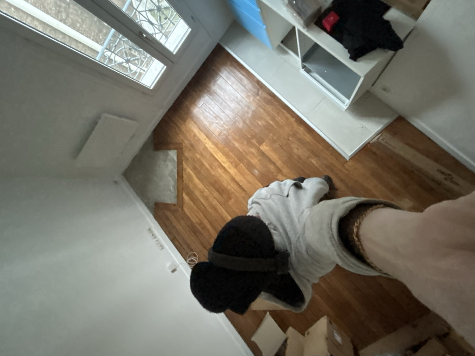 From a high angle, the photo shows a view into a room. The floor is wood paneling. Along the top right is a partially visible white shelf with items inside. A window is at the top left. At the bottom, a person extends their leg, clad in light grey pants and a black clog. Packing boxes are at the very bottom. The walls are white.
