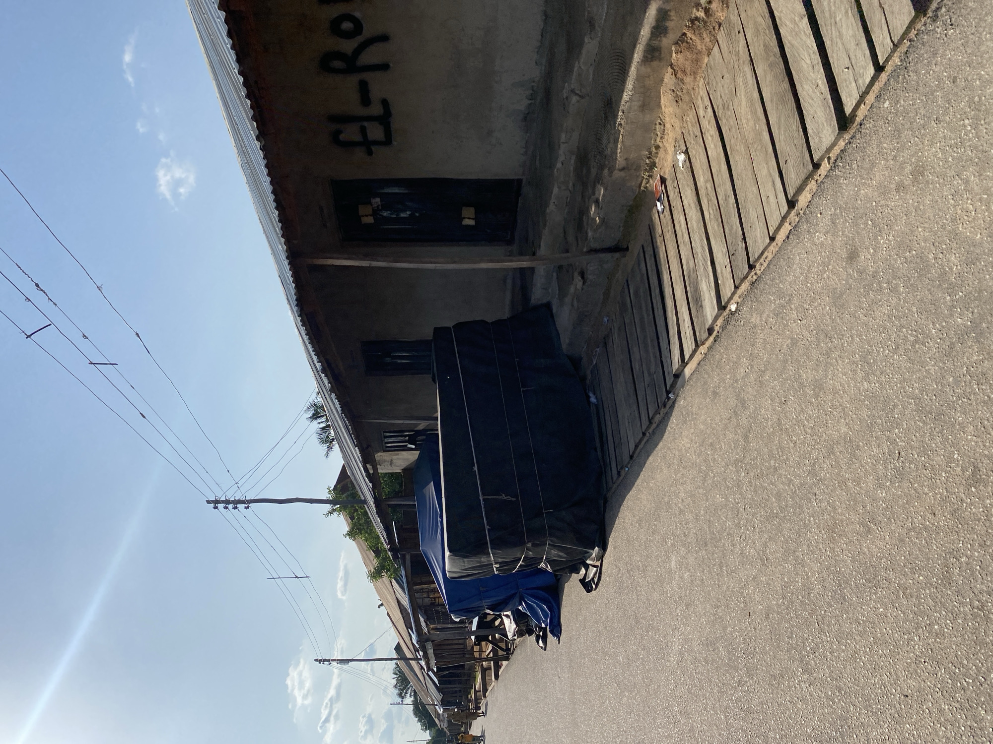 The image depicts a street scene in Jalingo, Nigeria, likely in the late afternoon given the long shadows. The perspective is from a low angle, looking up towards buildings and the sky. A paved road occupies the foreground, with a wooden boardwalk or structure running alongside it on the left. Beyond the boardwalk, a row of buildings is visible. These appear to be modest structures with corrugated metal roofs and concrete walls. Some of the buildings have openings covered with dark material, possibly tarpaulin or black plastic sheeting. One building has graffiti on its exterior wall, which appears to say "EL-RO".
The sky is a clear, bright blue with a few wispy clouds. Overhead, a network of power lines crisscrosses the sky, supported by wooden utility poles. Some of the buildings appear to be covered with blue and black tarpaulins, suggesting goods or structures are being protected from the elements. A few small figures are visible in the distance on the road, and some vegetation can be seen peeking out from behind the buildings. The overall impression is of a bustling, possibly commercial street in a town or city in Nigeria, with elements of everyday life and infrastructure clearly visible. The lighting suggests it is a sunny day.