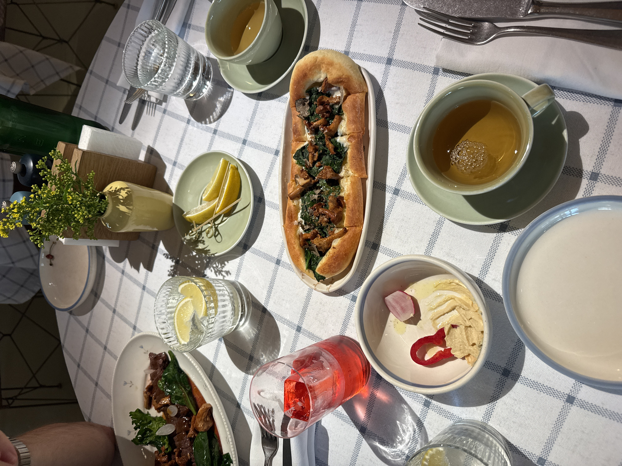 A table is set with a blue and white checkered tablecloth in what appears to be an indoor dining establishment. On the table, there are several dishes of food and drinks. In the foreground, a red beverage is in a glass with ice. To its right, there is a small white bowl with what looks like hummus, a slice of radish, and a piece of red pepper. Further to the right, a long, thin pastry with green vegetables and mushrooms is on a white plate. A cup of tea sits on a saucer to the right of the pastry. In the background, a plate of meat and greens is visible, as is a glass of water with lemon slices. A yellow vase with small yellow flowers stands near a wooden block and a green bottle. The lighting suggests it may be daytime, with sunlight casting shadows on the table. The presence of a fork and knife near the tea cup suggests a meal is in progress or about to begin. There is no visual information to ascertain the location as Moscow, Russia.