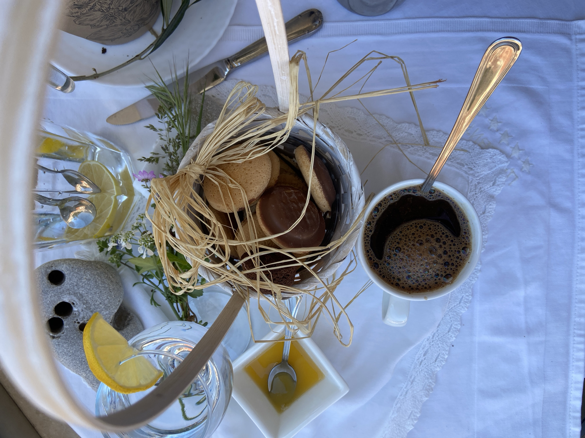 The image shows a table setting with a white lace-trimmed tablecloth. On the table are several items: a basket filled with cookies and decorated with raffia, a cup of black coffee with a spoon, a small square dish with honey and a spoon, a glass of water with a lemon slice, a stone with holes, a glass carafe filled with water and lemon slices, and a knife. Some greenery and flowers are also present. The image does not provide any information about the location in the city of Budva, Montenegro.