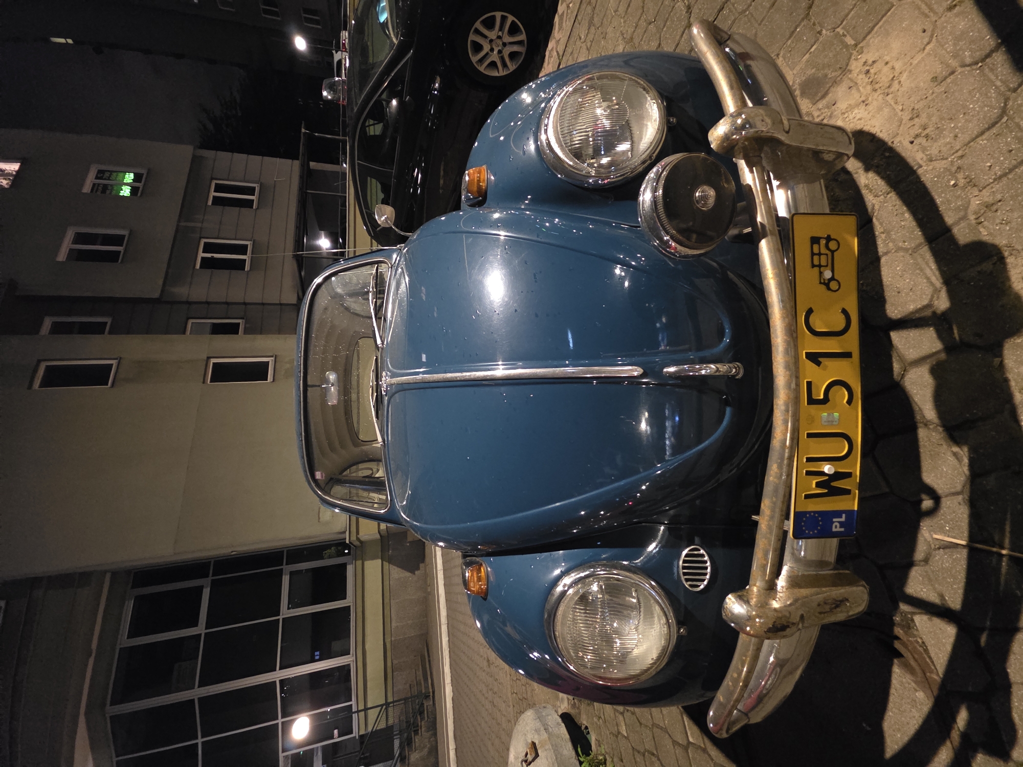 An extreme close-up, low-angle outdoor night photograph shows the front of a vintage, teal Volkswagen Beetle parked on a cobblestone street. The car is centrally framed, its iconic rounded hood and headlights dominating the foreground. The license plate, yellow with black lettering and European Union stars, reads "PL WU 51C" and features a small illustration of an old automobile. The car has a chrome bumper and a single, circular headlight mounted below the main headlights. To the right of the Beetle, a portion of a black modern sedan is visible. Behind the cars, a pale green, multi-story building with dark windows and some lights on is illuminated. The building appears to have architectural elements that suggest a city environment. A streetlight is visible to the left, casting a glow on the building's facade. The ground is paved with interlocking cobblestones. No people are present in the frame. The overall lighting is dim, typical of a nighttime urban setting.