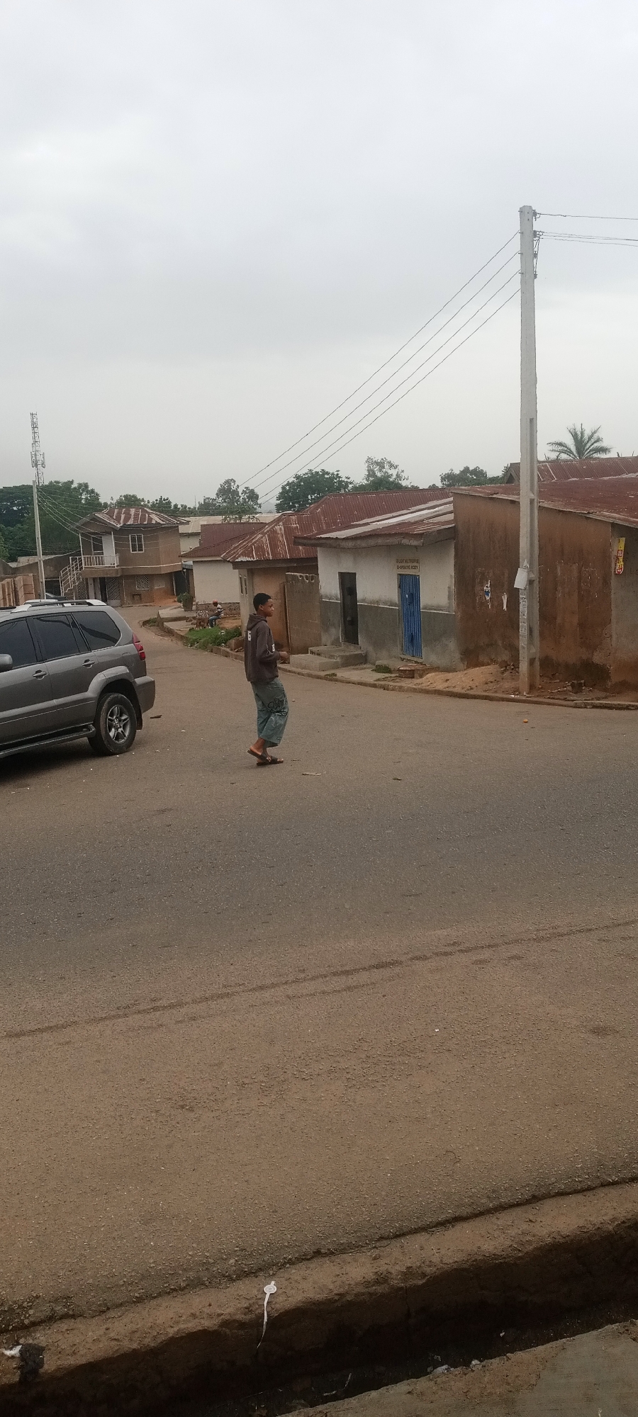 A residential street scene with a man crossing the road is depicted in the image. The man is wearing a dark hooded sweatshirt and light-colored patterned pants. He is also wearing sandals. A silver SUV is parked on the left side of the road, near the edge of the frame. The street is paved, and a narrow ditch runs along its edge at the bottom of the frame. Houses line the street, with varying architectural styles and materials, including corrugated metal roofs and painted walls. An overhead power line stretches across the scene, supported by a utility pole. The sky is overcast, suggesting a diffused light. The city or region is Unknown, Unknown.