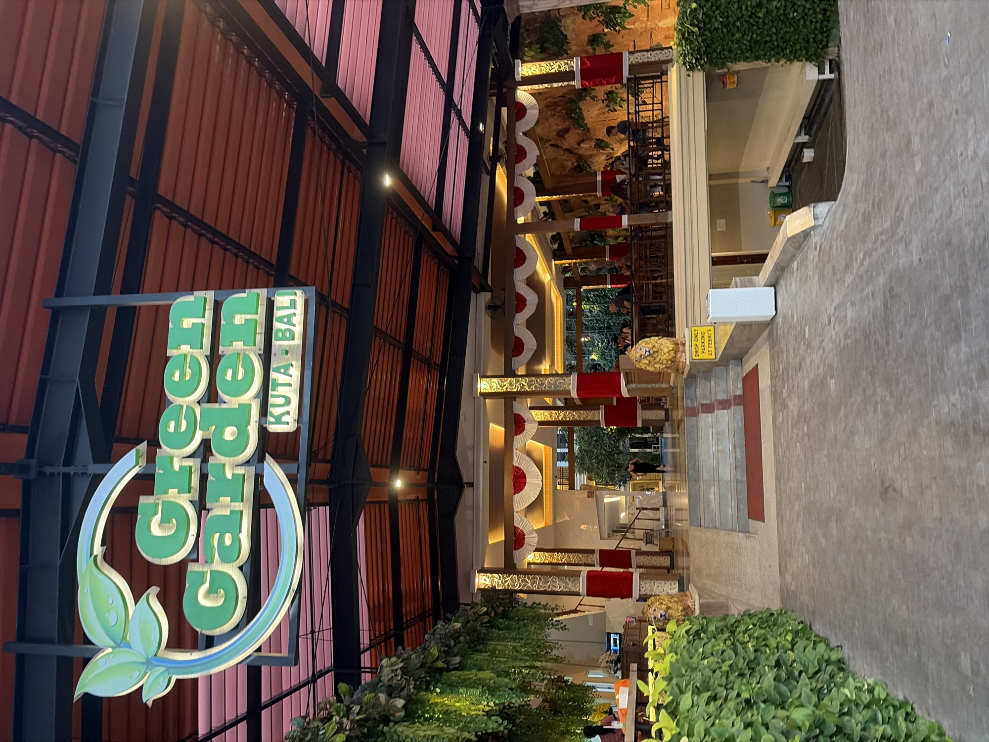 The image shows the exterior of a restaurant named "Green Garden" located in Kuta, Bali, Indonesia. The sign for the restaurant is prominent, featuring green lettering and a leaf motif. The building has a covered outdoor seating area with tables and chairs, where several patrons are seated. Decorative elements, including red and white banners and ornate pillars, are visible. Lush greenery, such as hanging plants and hedges, surrounds the entrance and pathways. A yellow sign with text that includes "DROP ONLY PARKING" is also present. In the background, there are stairs leading up to the seating area, and a man is walking through an open doorway. The lighting suggests it is evening or late afternoon.