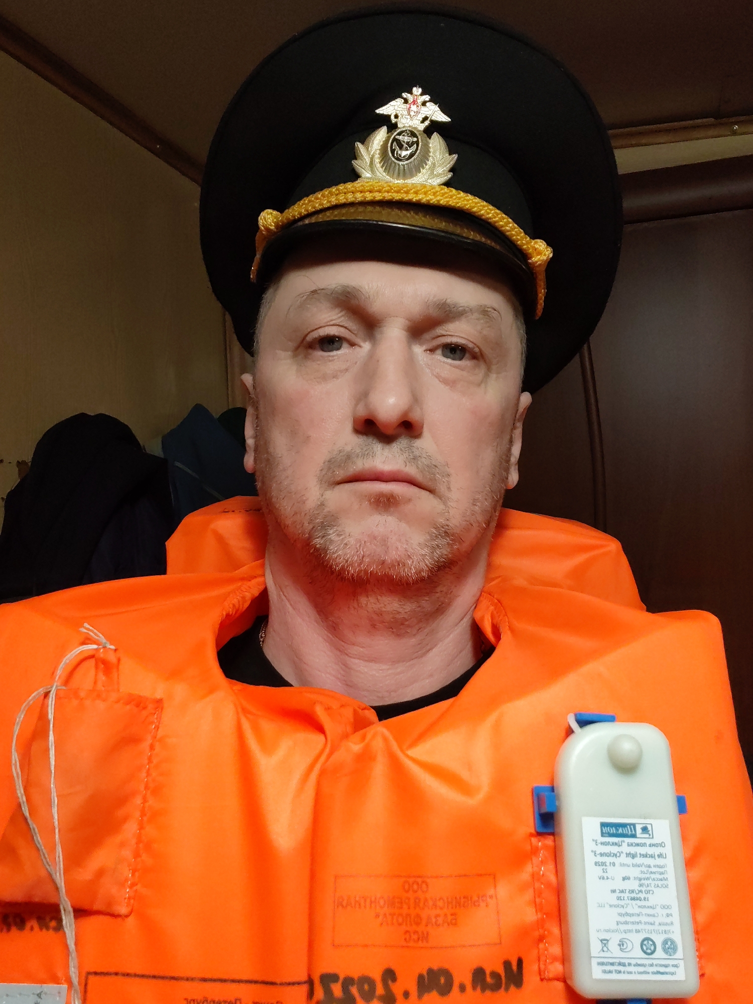 A man is wearing a black hat with a gold emblem and a bright orange life vest. The man is facing the camera. The text on the life vest is in Russian. The location is indoors. The city of Rybinsk, Russia cannot be inferred from this image.