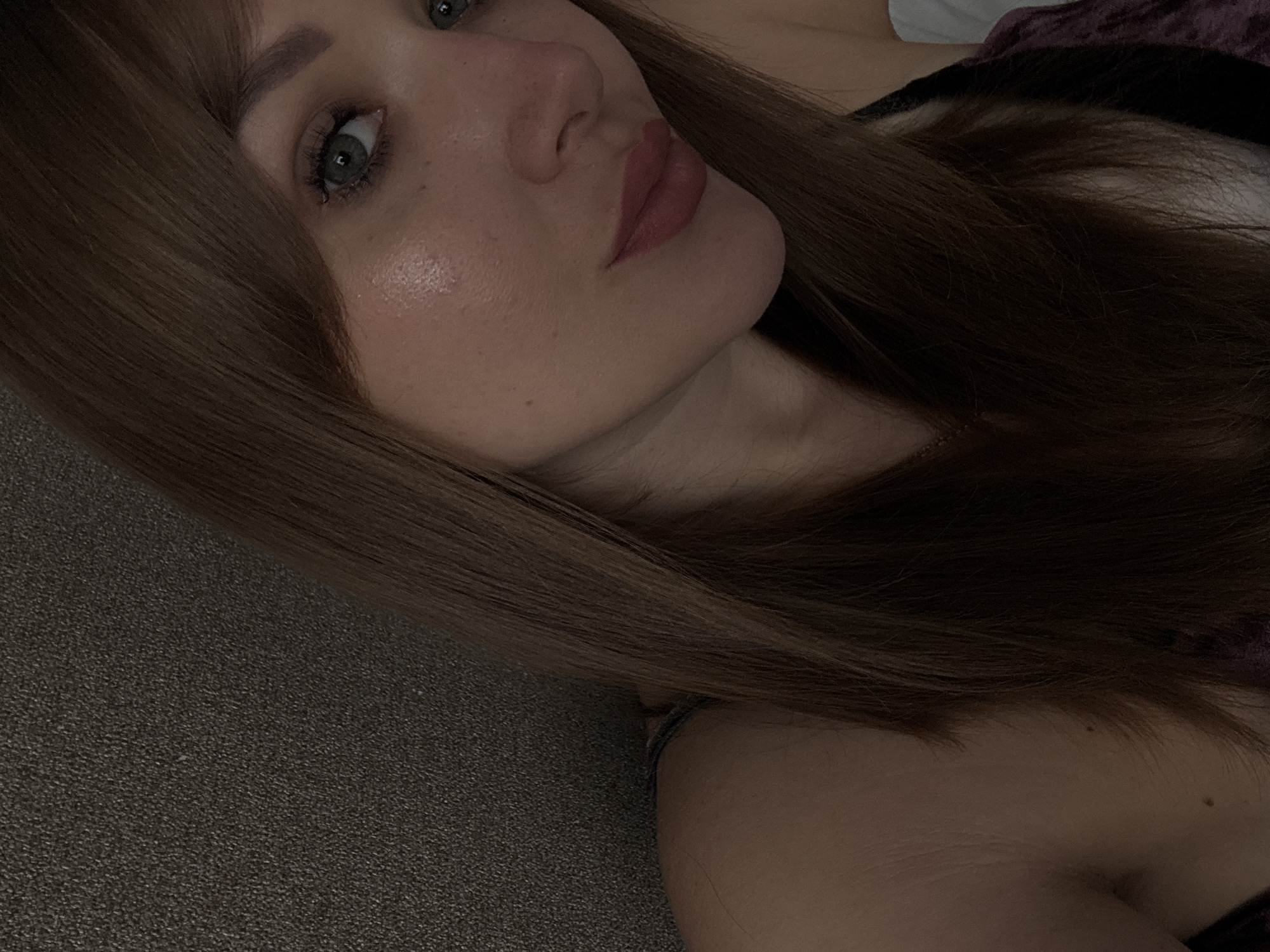 A close-up, oblique-angle shot depicts a young woman with light skin and light brown hair. Her face is the main focus, featuring green eyes and pink lipstick. The left side of her face is illuminated, showing skin texture and freckles. Some of her hair is in the foreground to the left and is out of focus. A dark, textured surface, possibly a couch, fills the lower left portion of the image. Part of her shoulder is visible in the lower right. The top right shows hints of fabric, possibly clothing or a blanket. No specific details point to Chaikovsky, Russia, or any other location.