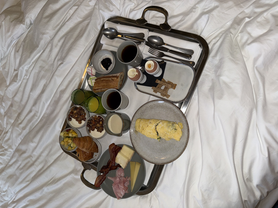 A rectangular metallic breakfast tray with brass-colored handles is centrally positioned on a white, wrinkled bedspread. The tray is fully occupied with various breakfast items. From left to right: a small bowl contains diced fruit (pineapple, mango, melon) next to two croissants. Below these, a gray plate holds slices of cured ham, two types of cheese, and several strips of bacon. Two small bowls of yogurt topped with granola are present, alongside a pitcher of cream or milk and two cups of black coffee. Two glasses contain beverages, one orange juice and one green juice. A section of baguette bread is accompanied by two pats of butter and a small bowl of dark jam. A separate gray plate holds a rolled omelet. A small rectangular white tray features two soft-boiled eggs in white egg cups, positioned next to a stack of small rectangular crackers arranged in a hashtag pattern. Multiple sets of silverware, including forks, knives, and spoons, are neatly arranged on the right side of the main tray, wrapped in white napkins. No individuals are present. The scene depicts an indoor setting, likely a hotel room, but the exact location within Paris, France, cannot be determined from the visual evidence.