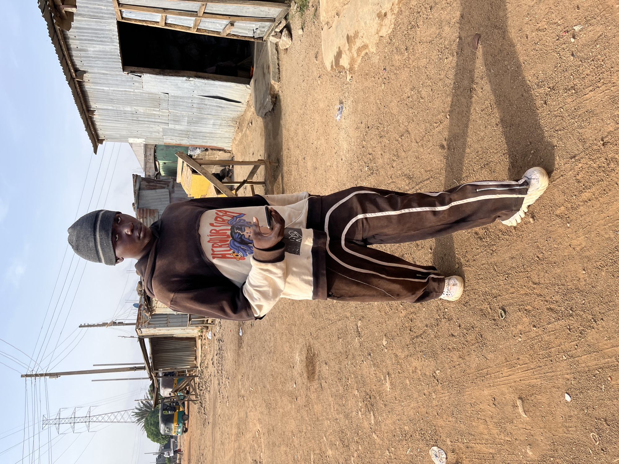 A young Black man stands in a dirt road in Jos, Nigeria, wearing a brown tracksuit with white stripes and a grey beanie. He holds a phone in his hand, facing the camera. Behind him is a corrugated metal structure, indicative of informal construction. Power lines and utility poles are visible in the background. Other vehicles and structures are partially visible further down the road.