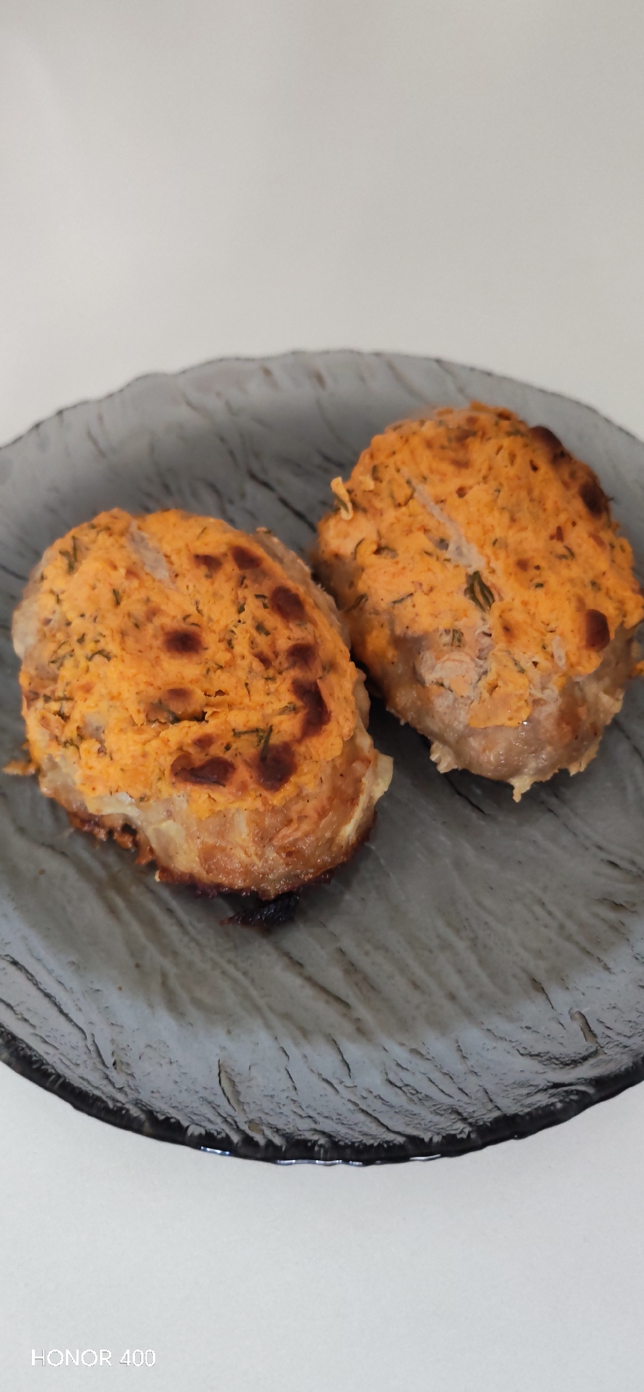Two cooked, round meat patties with an orange topping are presented on a gray, textured glass plate. The environment appears to be indoors, with a white background. The location information provided is not evident in the image. No people are visible.