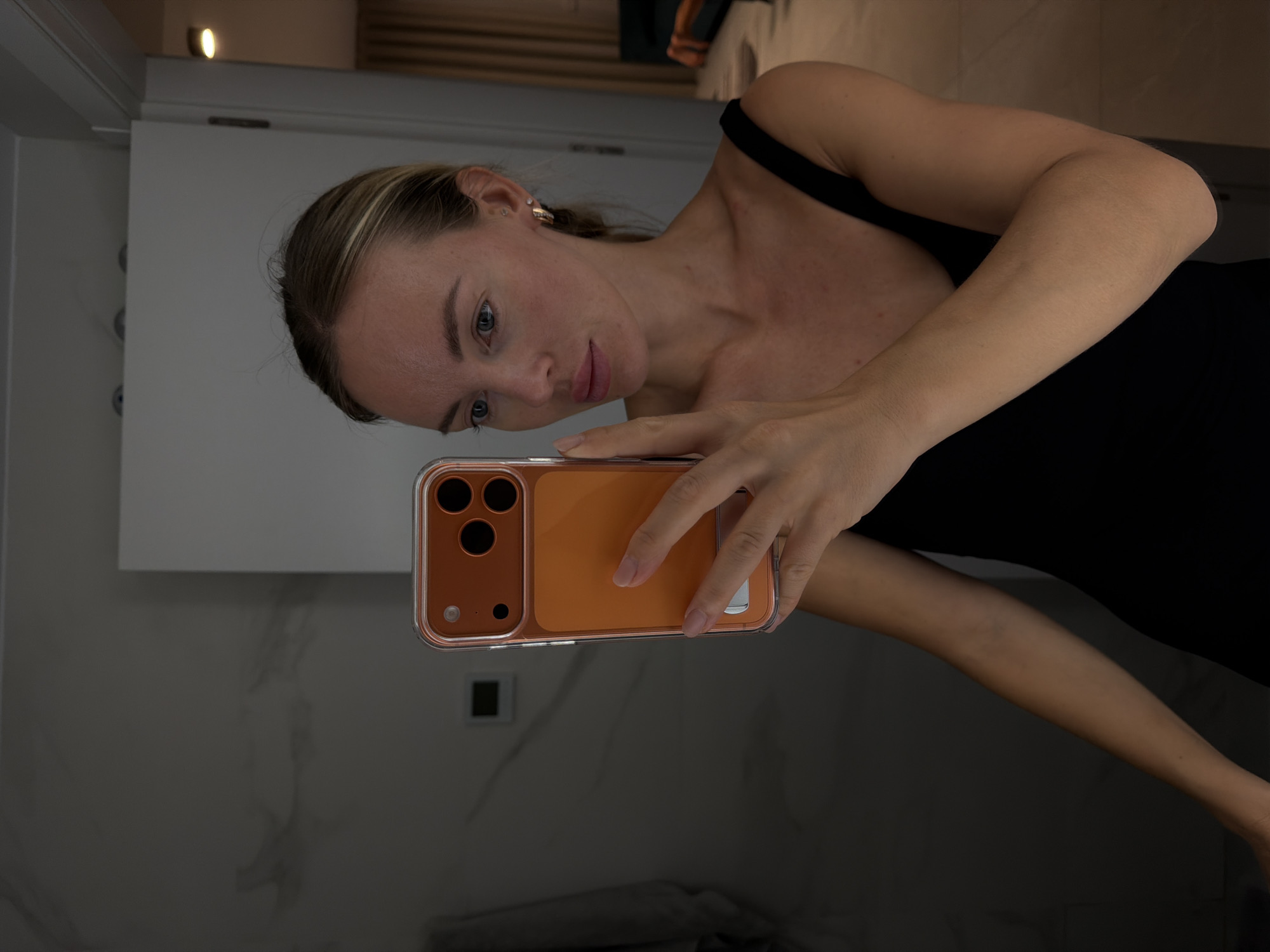 In Finestrat, Spain, a young woman with blonde hair pulled back and blue eyes takes a mirror selfie. She is wearing a black tank top and gold hoop earrings. Her right hand holds an orange-colored phone case with multiple camera lenses and a silver kickstand. She is in a bathroom with marbled walls and a white door in the background. A towel hangs on the left side of the image.