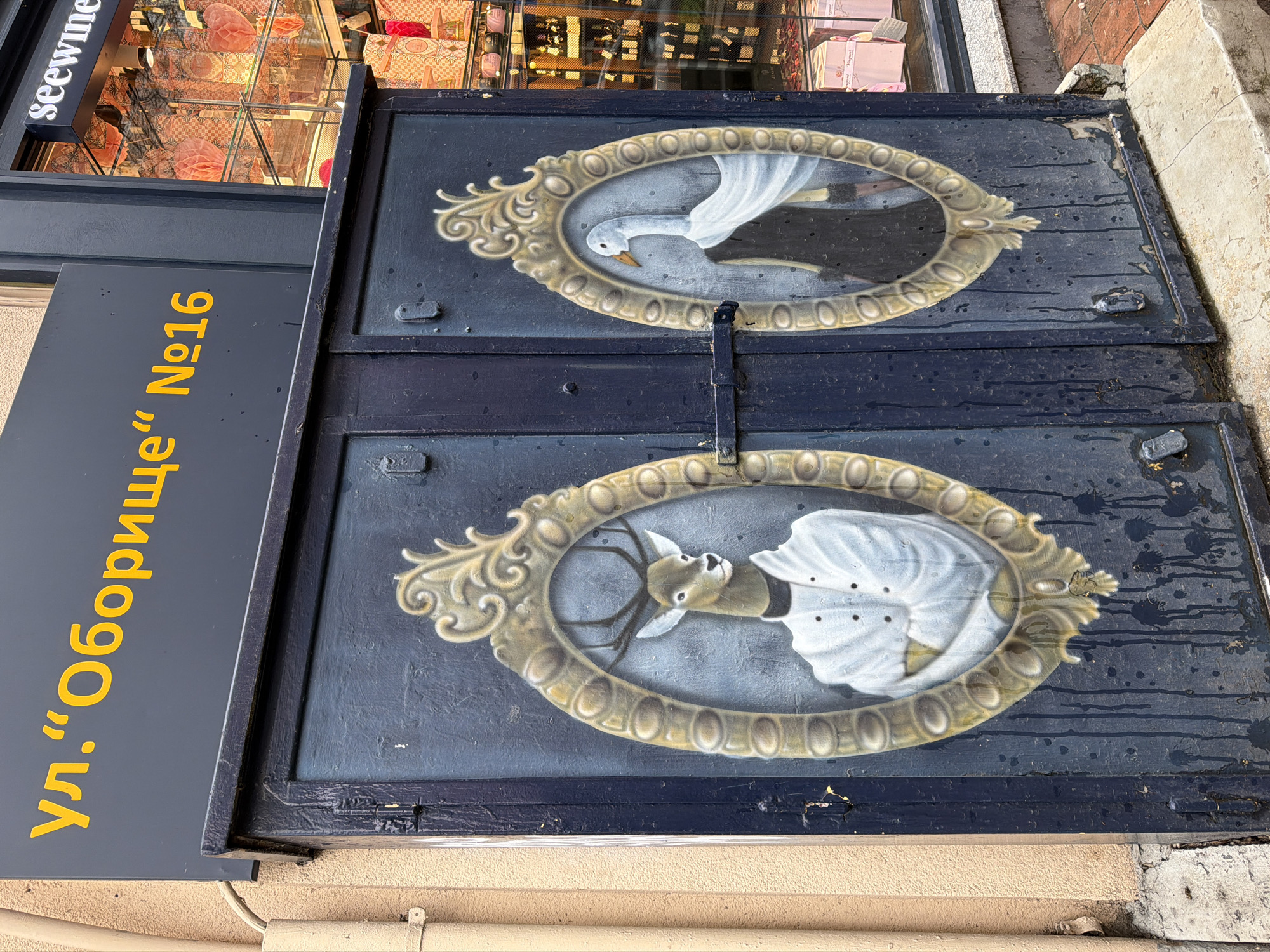 Two dark blue metal utility cabinet doors, adorned with anthropomorphic animal portraits, are situated on "ул. "Оборище" №16" (Oborishte Street No. 16) in Sofia, Bulgaria. The left door features a deer with antlers, rendered in a white chef's coat, depicted within an ornate gold-framed oval. The deer's forelegs are crossed over its chest. The right door displays a white goose with an orange beak, dressed in a dark garment with a flowing white shawl or cape, also within a similar ornate gold-framed oval. A horizontal metal latch secures the two doors together, positioned between the two painted portraits. Above the doors, a dark grey sign with yellow Cyrillic text reads "ул. "Оборище" №16". To the immediate right, a glass storefront is visible, containing merchandise, including red honeycomb decorations and various packaged goods, with part of a "seewine" sign visible at the top. To the left, a beige stucco wall with a light-colored conduit pipe runs vertically. The cabinet doors show signs of wear, including vertical dark streaks consistent with water runoff and some paint chipping, particularly towards the bottom. The ground directly beneath consists of a concrete slab, transitioning to brick paving further to the right.
