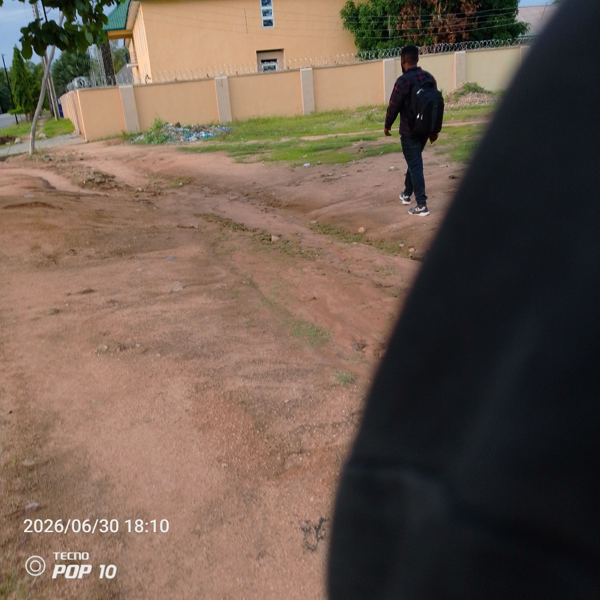 A person walks away from the camera on a dirt path. The person is wearing a red and black plaid shirt, dark pants, and athletic shoes. They have a black backpack on. The path is flanked by dry, sandy soil with sparse patches of green grass. In the background, there is a light yellow building with a green roof and barbed wire along the top of a cream-colored wall. Some trees and bushes are visible behind the wall. There is also some litter on the ground near the wall. The sky appears overcast. The bottom of the image contains visible text. The date "2026/06/30 18:10" is displayed, along with the branding "TECNO POP 10". The scene appears to be outdoors in a semi-urban or rural area, possibly in Jalingo, Nigeria, given the provided location context. The time of day is late afternoon or early evening. The overall mood is casual and everyday.
