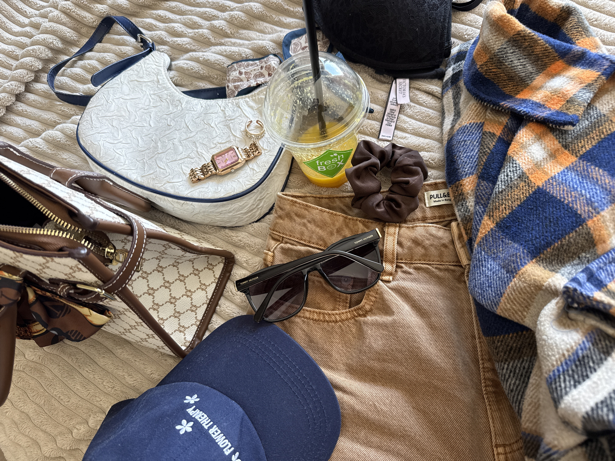 An overhead view depicts an arrangement of personal items and apparel laid out on a light beige, textured surface. In the upper left, a dark blue baseball cap with white "FLOWER THERAPY" embroidery and two flower icons is visible, alongside a beige and brown patterned handbag featuring brown leather-like trim, a gold-tone zipper, and a brown patterned silk scarf tied to its handle. To the right, a white crescent-shaped bag with a textured surface and blue trim holds a gold-tone rectangular wristwatch with a pink face and a gold-tone ring. A pair of tan denim trousers, bearing a "PULL&BEAR" label, are laid across the lower center, with black "TOMMY HILFIGER" sunglasses resting on them. A clear plastic cup containing an orange liquid, a black straw, and a green "Fresh Box" label is positioned near a brown scrunchie. A black lace bralette with a visible "VICTORIA'S SECRET" label is situated towards the right. A folded plaid scarf, featuring blue, orange, white, and grey stripes, occupies the bottom of the frame. The scene is located in the city of Plovdiv, Bulgaria.