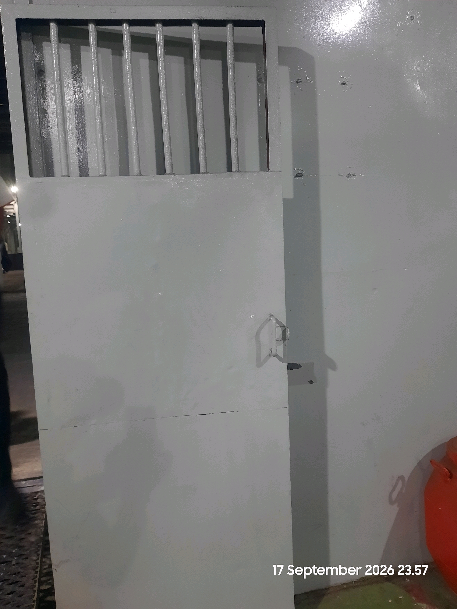 The image shows a metal door with a barred window at the top. The door is painted light gray and has a simple metal handle. The window appears to be set into the upper portion of the door, with vertical bars providing security. The wall to the right of the door is also painted gray and shows some minor imperfections, such as small dents or scratches. A bright orange object, possibly a container or piece of equipment, is visible in the lower right corner of the image. The overall impression is of an interior space that might be utilitarian or secure, such as a storage area, a utility room, or a part of a building with controlled access. The text overlay indicates the date and time "17 September 2026 23.57". No specific location in Gending, Indonesia is discernible from the visual elements of the image.