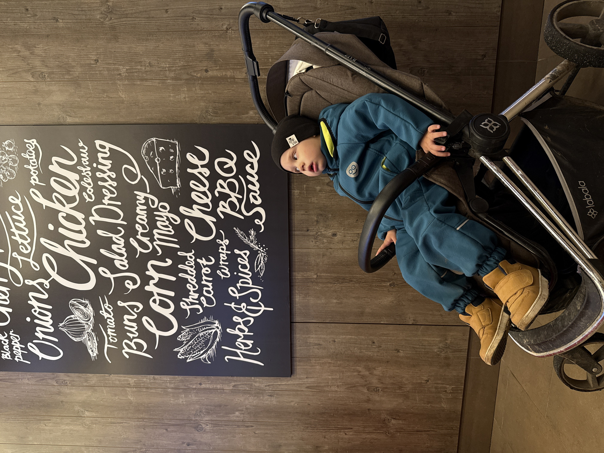 This photograph, taken in Rusanovo, Russia, is presented rotated 90 degrees counter-clockwise, showing a young child seated upright in a stroller within an indoor setting. The child, likely a toddler, wears a blue full-body suit or overall, a black beanie with a small white tag, and light brown boots. They appear calm and observant, looking slightly towards the right of the frame.
The dark-colored stroller, bearing the brand "labala" on its side and with a bag marked "RAIZ" attached to its handle, is positioned on what appears to be a wooden floor or wall with horizontal planks. To the child's left is a dark rectangular board, likely a menu or ingredient list, featuring various food items and drawings in white, chalk-style lettering. Visible text on the board includes "Black pepper", "Onions", "Lettuce", "potatoes", "Chicken", "Tomato", "Buns", "Salad Dressing", "Creamy", "Mayo", "Corn", "Shredded Carrot", "Cheese", "wraps", "BBQ", "Herbs & Spices", and "& Sauce". Illustrations of onions, a corn cob, and a block of cheese accompany the text. The overall scene suggests a casual commercial establishment such as a cafe or restaurant.