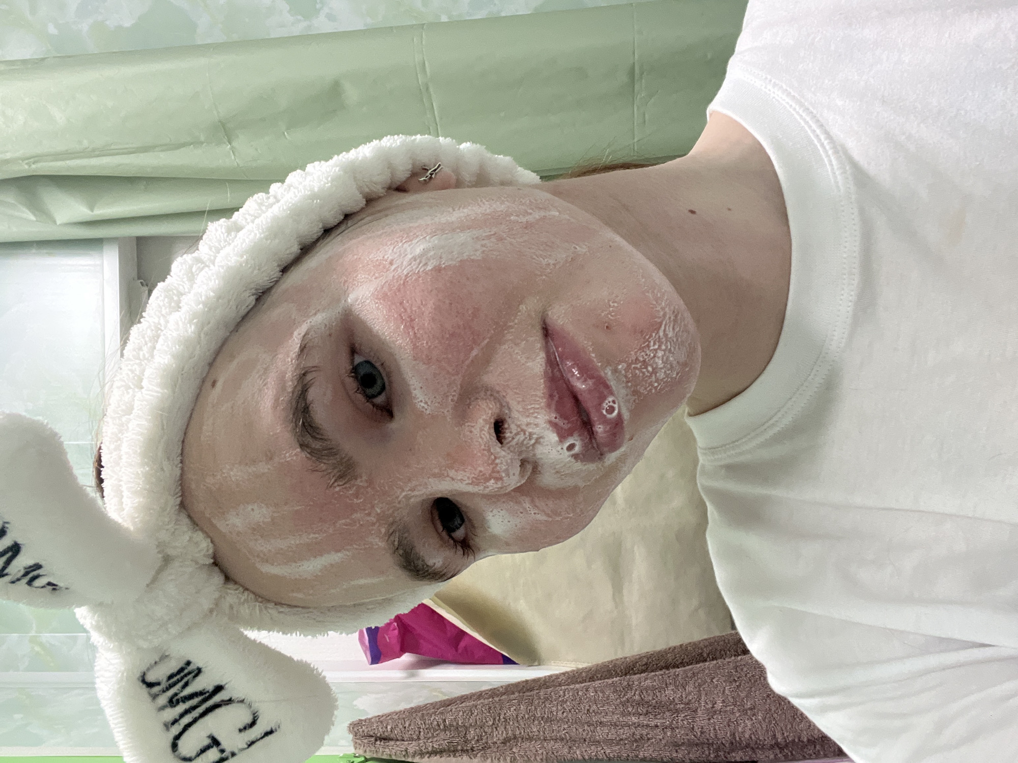 A person with a white facial mask is centered in the frame, wearing a white plush headband with the letters "OMG" on it. Their face is tilted towards the camera. They are wearing a white t-shirt. The background consists of a light green surface and a window. A brown towel and a pink object are visible on a surface below.
It is not possible to confirm the location in the city of Chaikovsky, Russia.