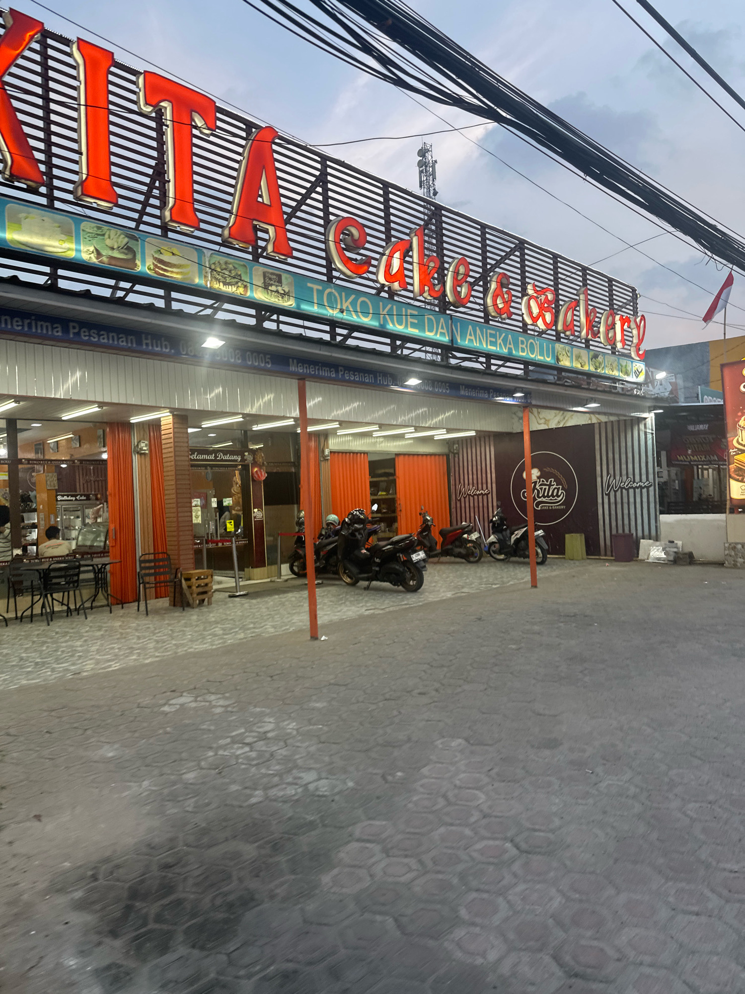 The image shows the exterior of a bakery and cake shop called "Kita Cake & Bakery" located in Saruni, Indonesia. The shop has a prominent red neon sign with the name of the establishment. Below the sign, there is a display of various cakes and pastries. The entrance to the shop features orange folding doors and a "Selamat Datang" (Welcome) sign. Several motorcycles are parked in front of the shop, and some tables and chairs are set up outside. The pavement in front of the shop is made of hexagonal concrete blocks. The sky is overcast, suggesting it might be dusk or an overcast day. There is an Indonesian flag displayed on a pole to the right of the building.