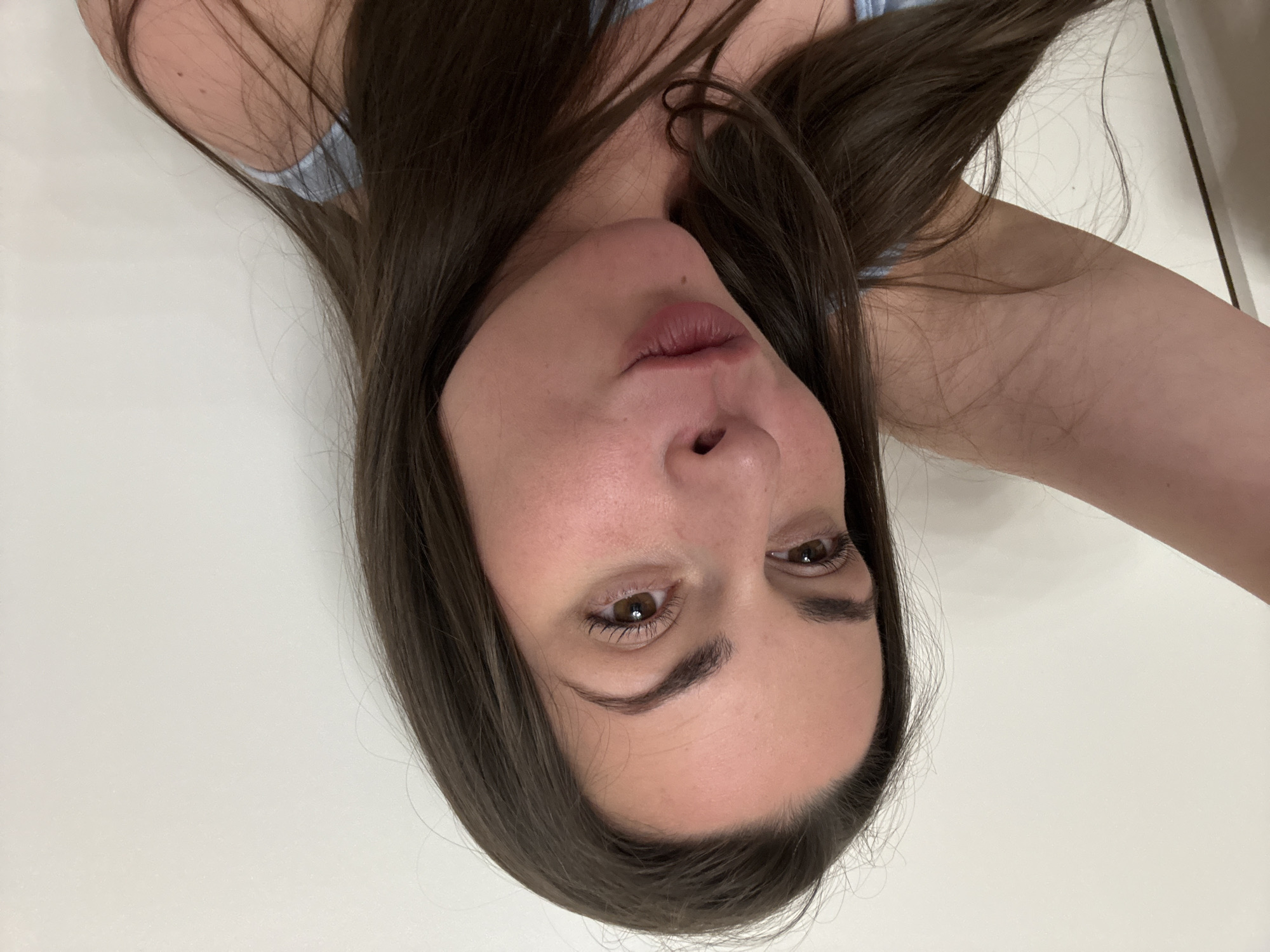 A woman with long, dark brown hair is taking a selfie against a white wall. She is looking to her left, with her lips pursed. Her right arm is raised and bent, with her hand out of frame. She is wearing a light blue top.