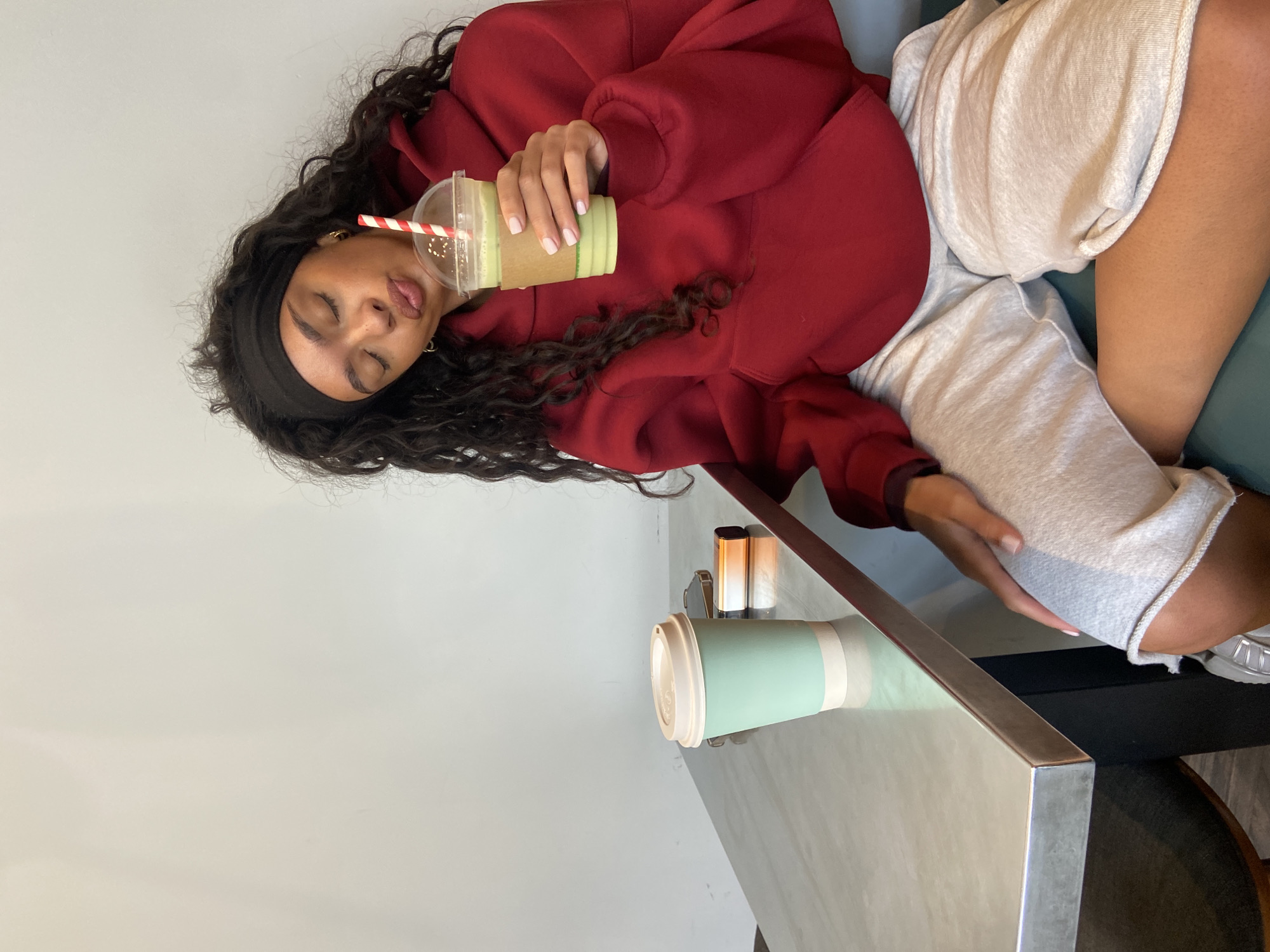 A woman with long, dark curly hair and a black headband sits at a table indoors. She is wearing a red hoodie and grey shorts, holding a green smoothie with a red and white striped straw. Her eyes are closed. On the table, there is a light green coffee cup and some beauty products. The scene is likely in a cafe in London, United Kingdom, given the style of the furniture and beverages.